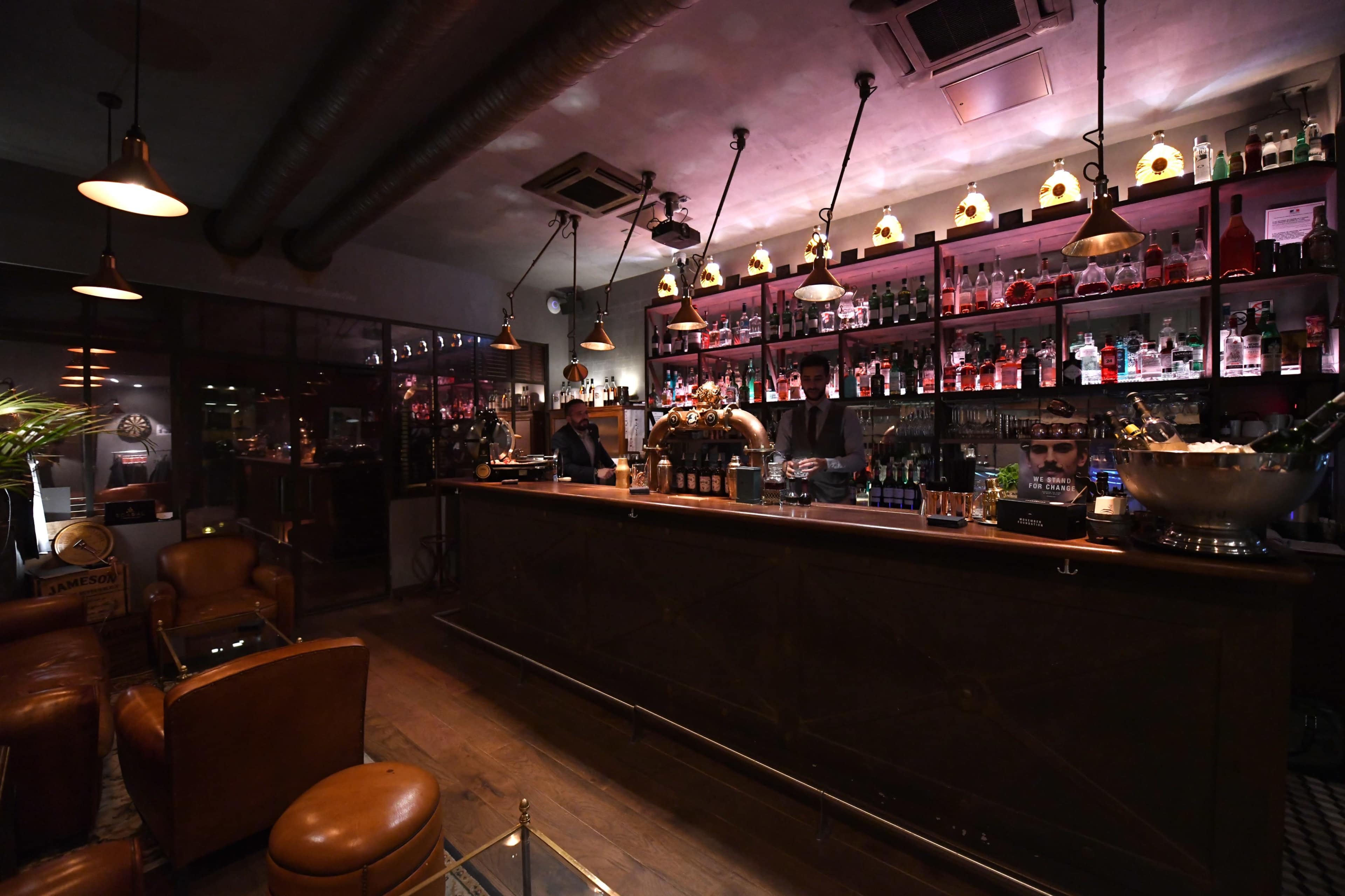 A dimly lit bar with a long wooden counter, shelves filled with various bottles, and leather chairs arranged around the space.