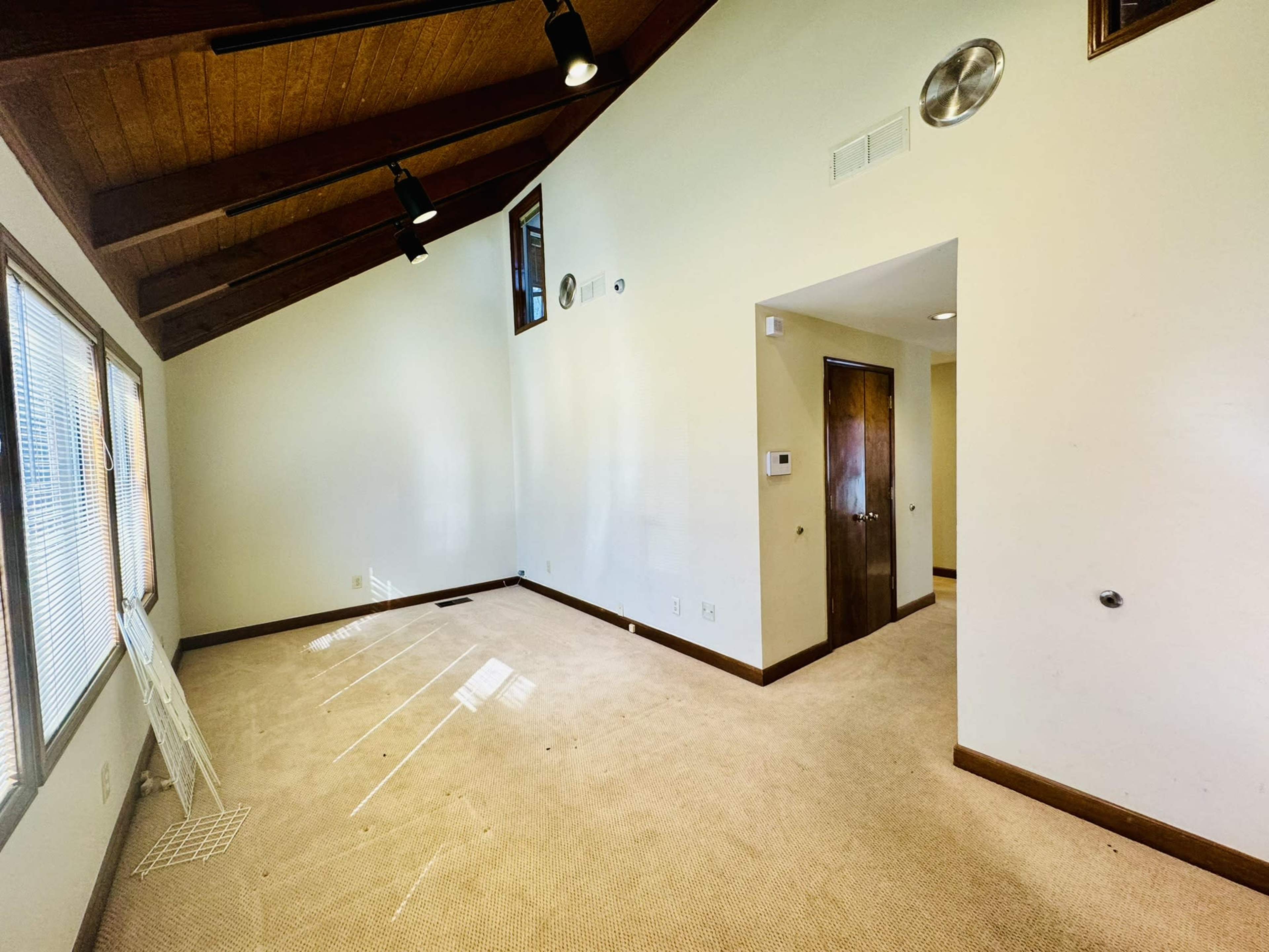 A spacious room with a high, sloped ceiling and carpeted flooring, featuring windows along one wall and an opening leading to another area.