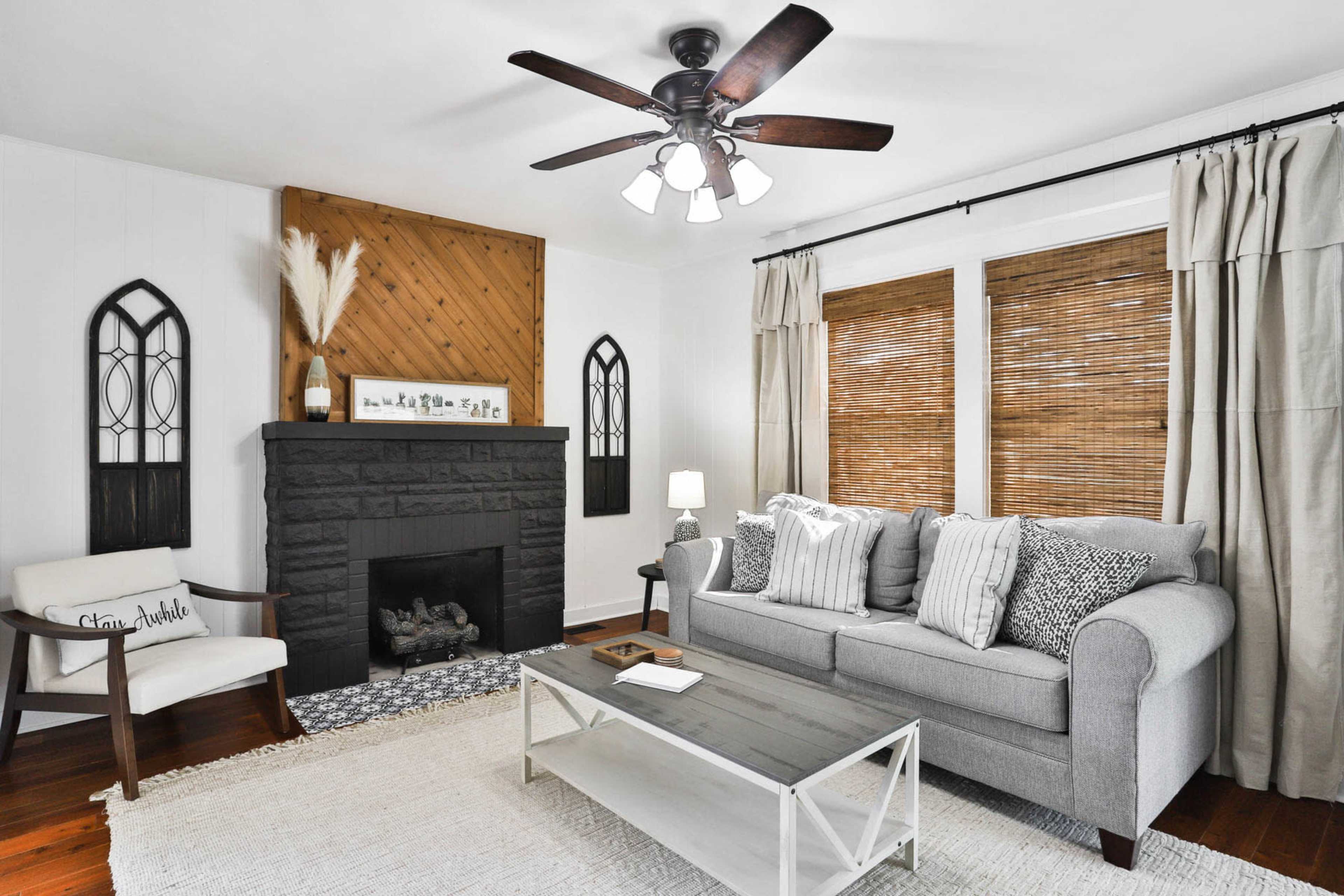 A styled living room features a gray sofa with patterned cushions, a white coffee table, and a black stone fireplace, accented by bamboo shades and decorative wall elements.