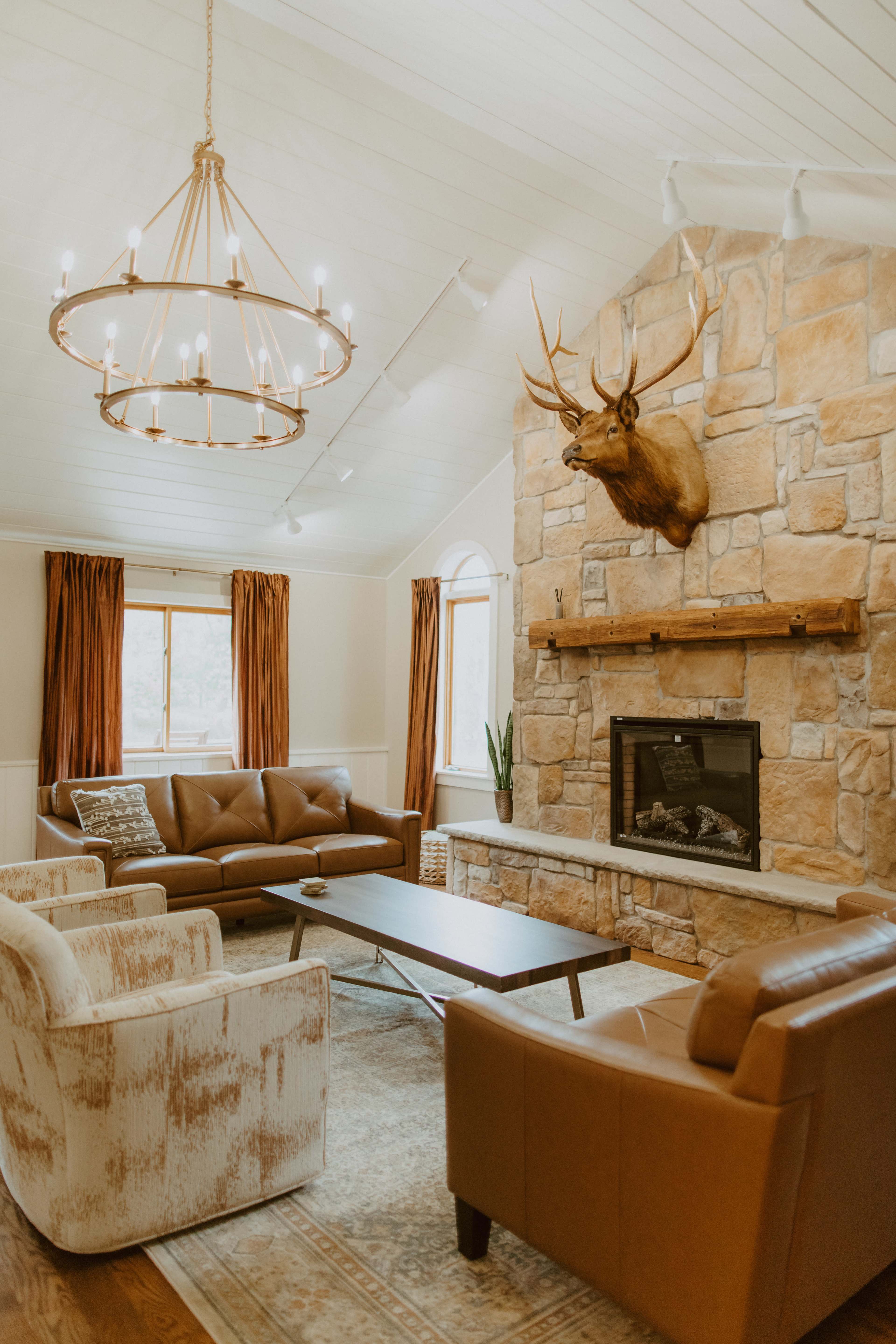 The living room features a stone fireplace with a mounted deer head, surrounded by a brown sofa, two patterned armchairs, a coffee table, and large windows with curtains.