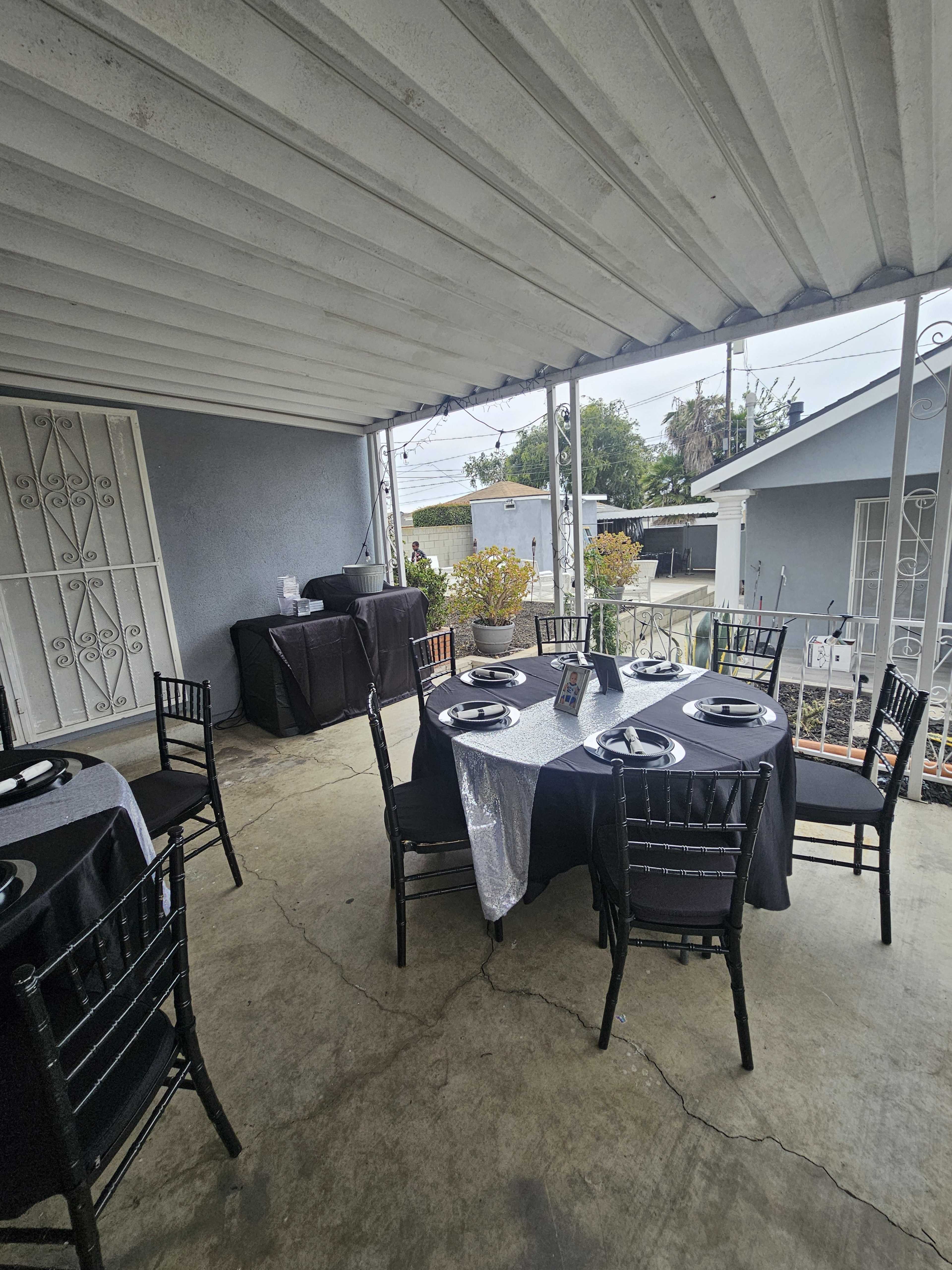 A covered outdoor area features two round tables set with black tablecloths and plates, alongside a grill and potted plants.