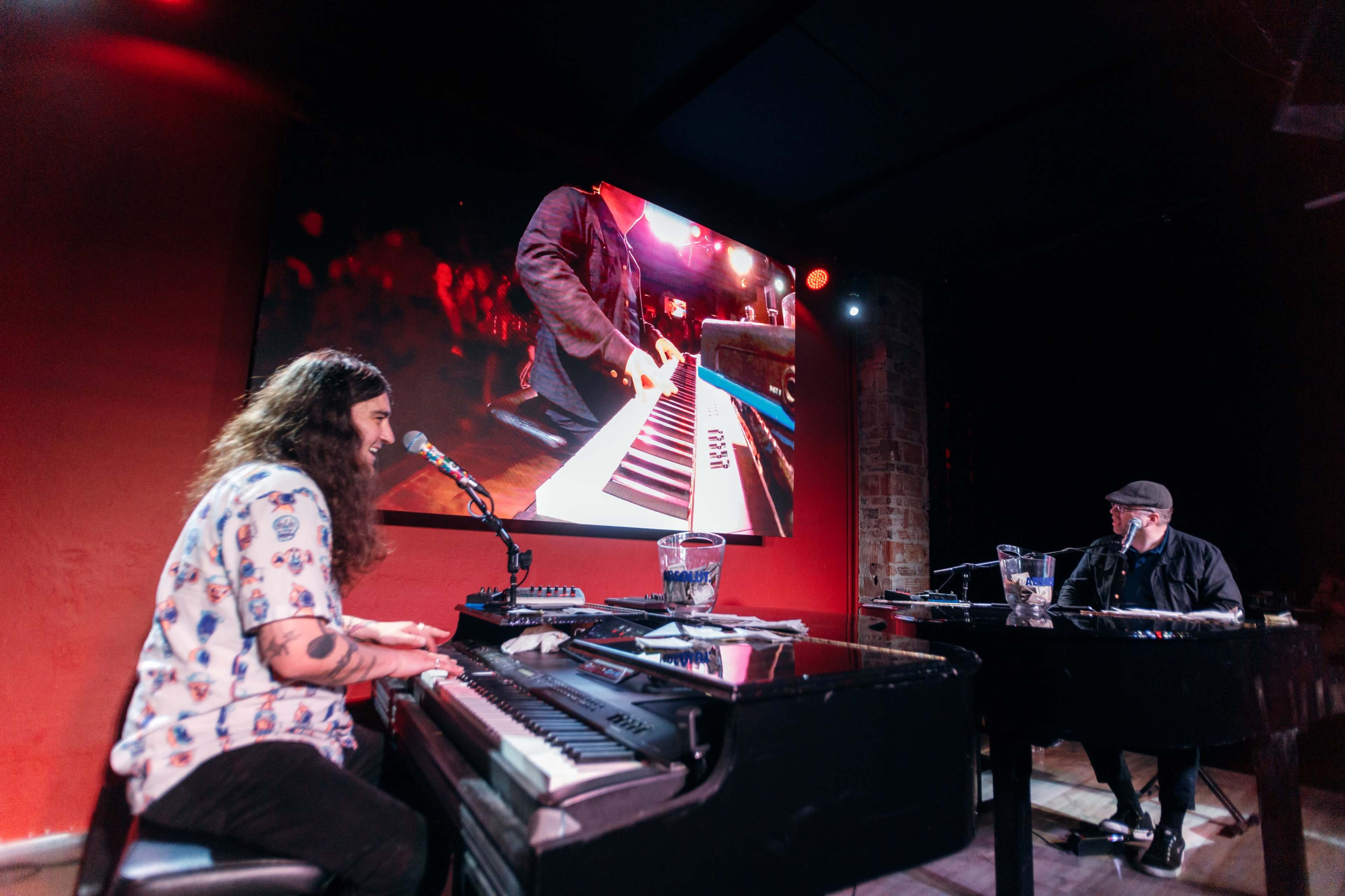 Two musicians perform at pianos on stage, with one playing and singing into a microphone while the other observes, and a video screen displays a close-up of the playing hands.