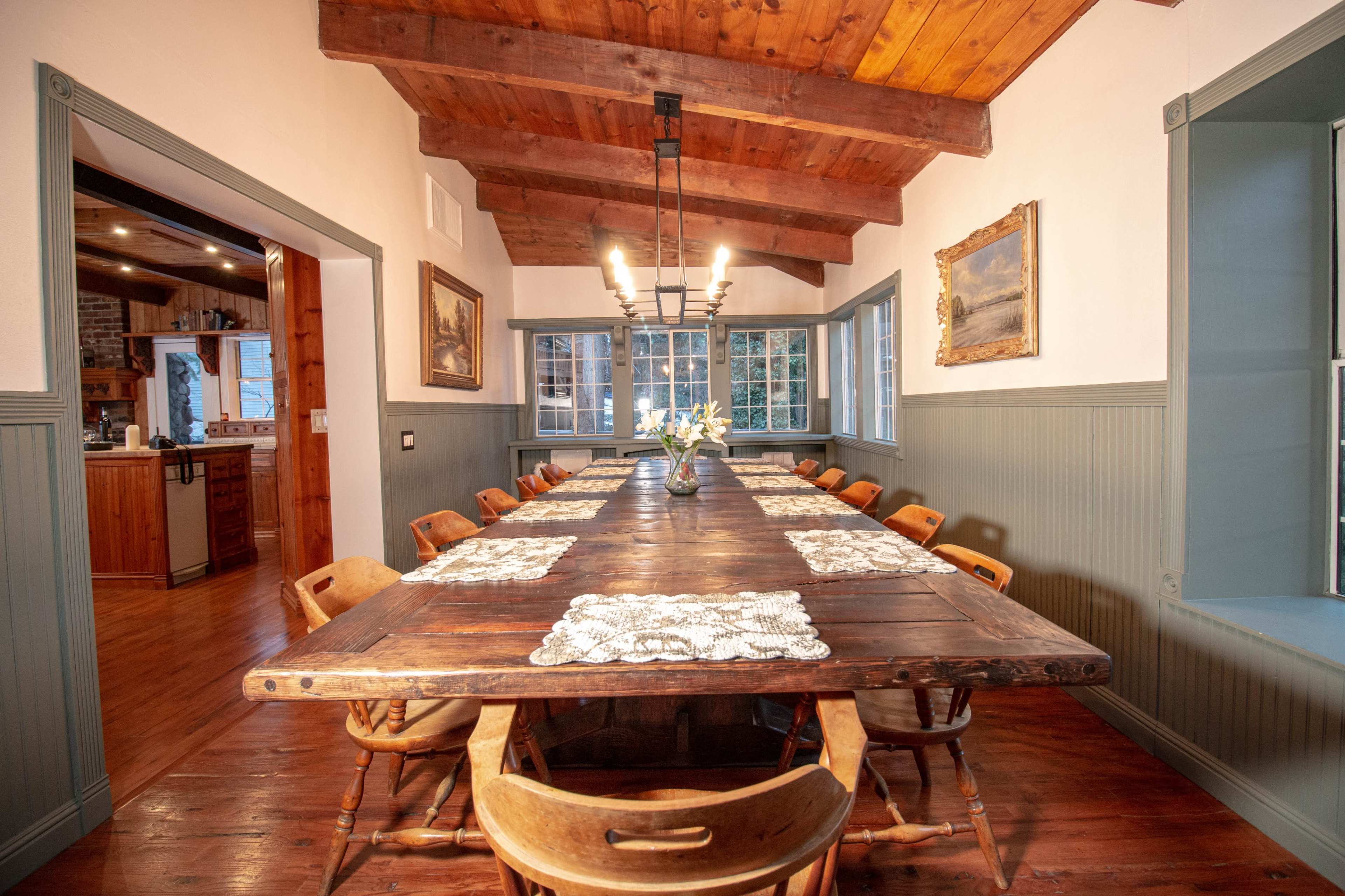 A long wooden dining table is set in a room with high, beamed ceilings and large windows, flanked by rustic chairs and decorated with a simple flower vase.