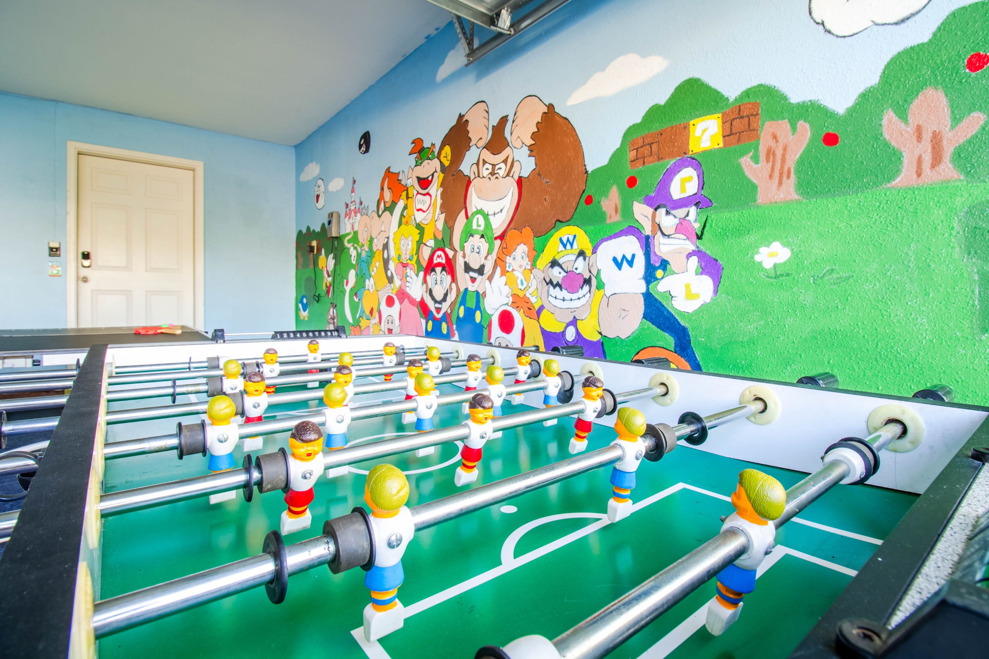 A foosball table with figurines is set against a colorful mural depicting video game characters in a game room.