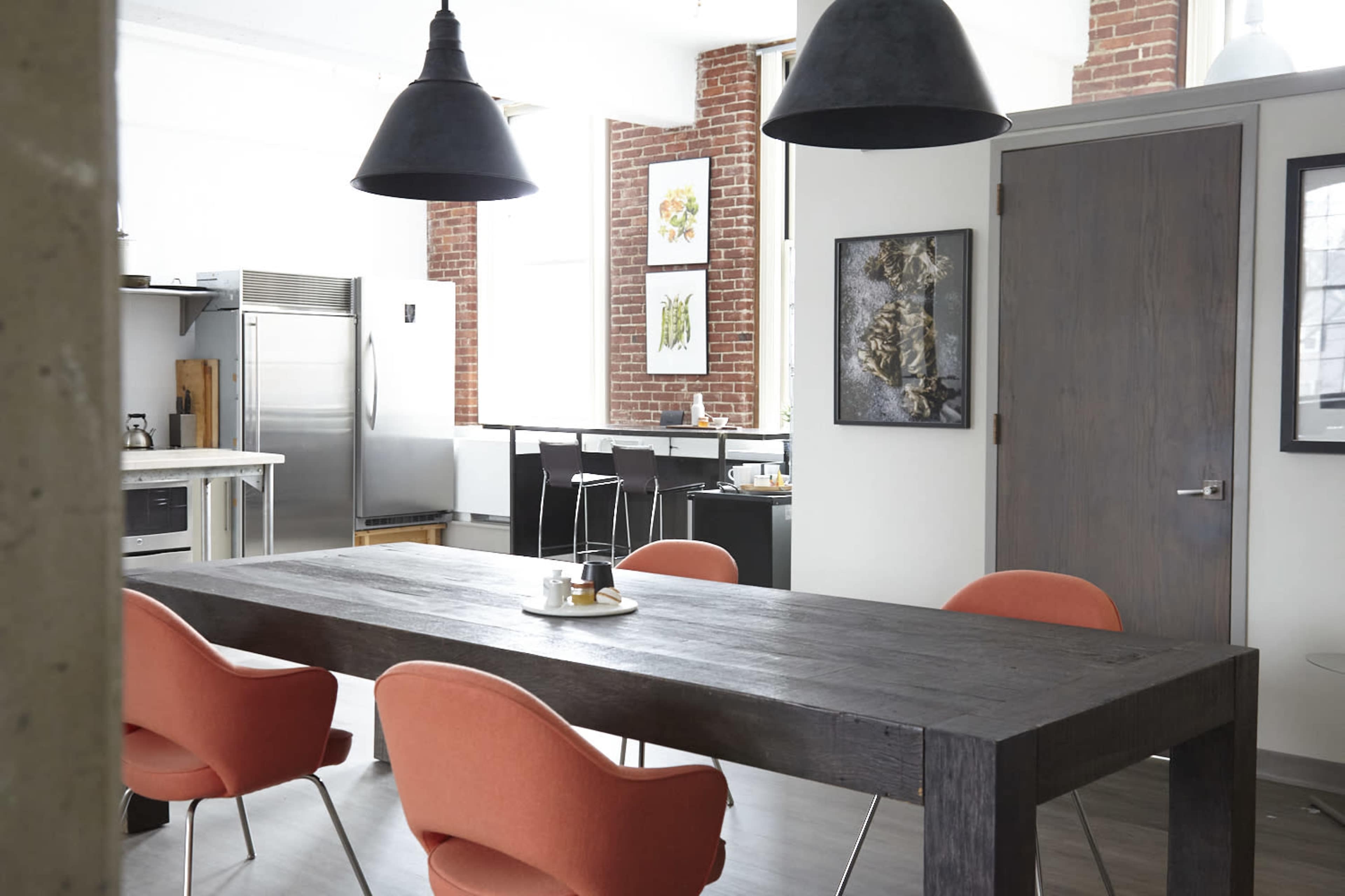 A modern kitchen and dining area featuring a wooden table with orange chairs, a stainless steel refrigerator, and pendant lighting.