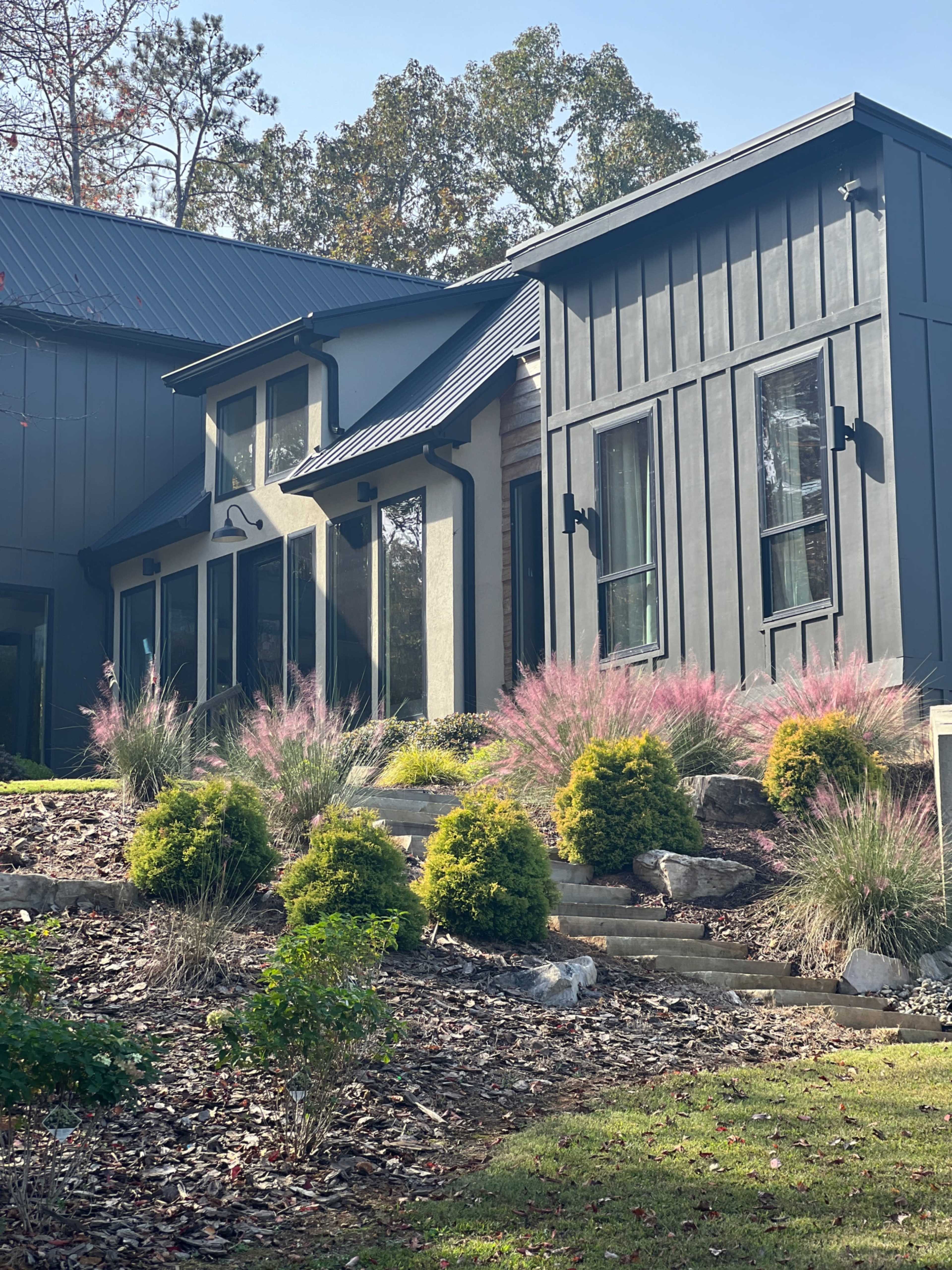 The image shows a modern house with a dark exterior, surrounded by landscaped gardens featuring ornamental grasses and shrubs.