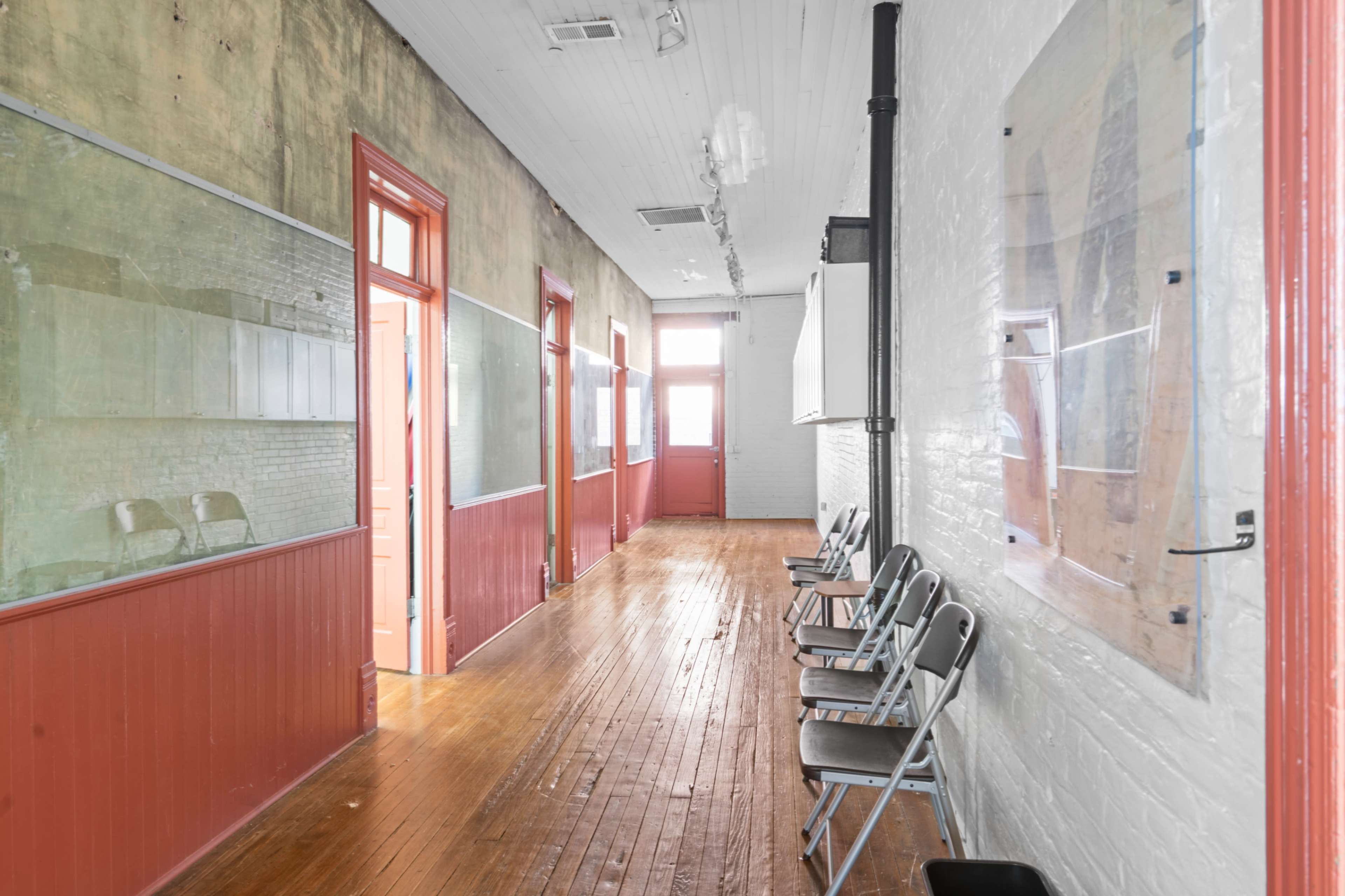 The image shows a narrow hallway with wooden floors, red-painted walls, and several folding chairs lined up against one side.