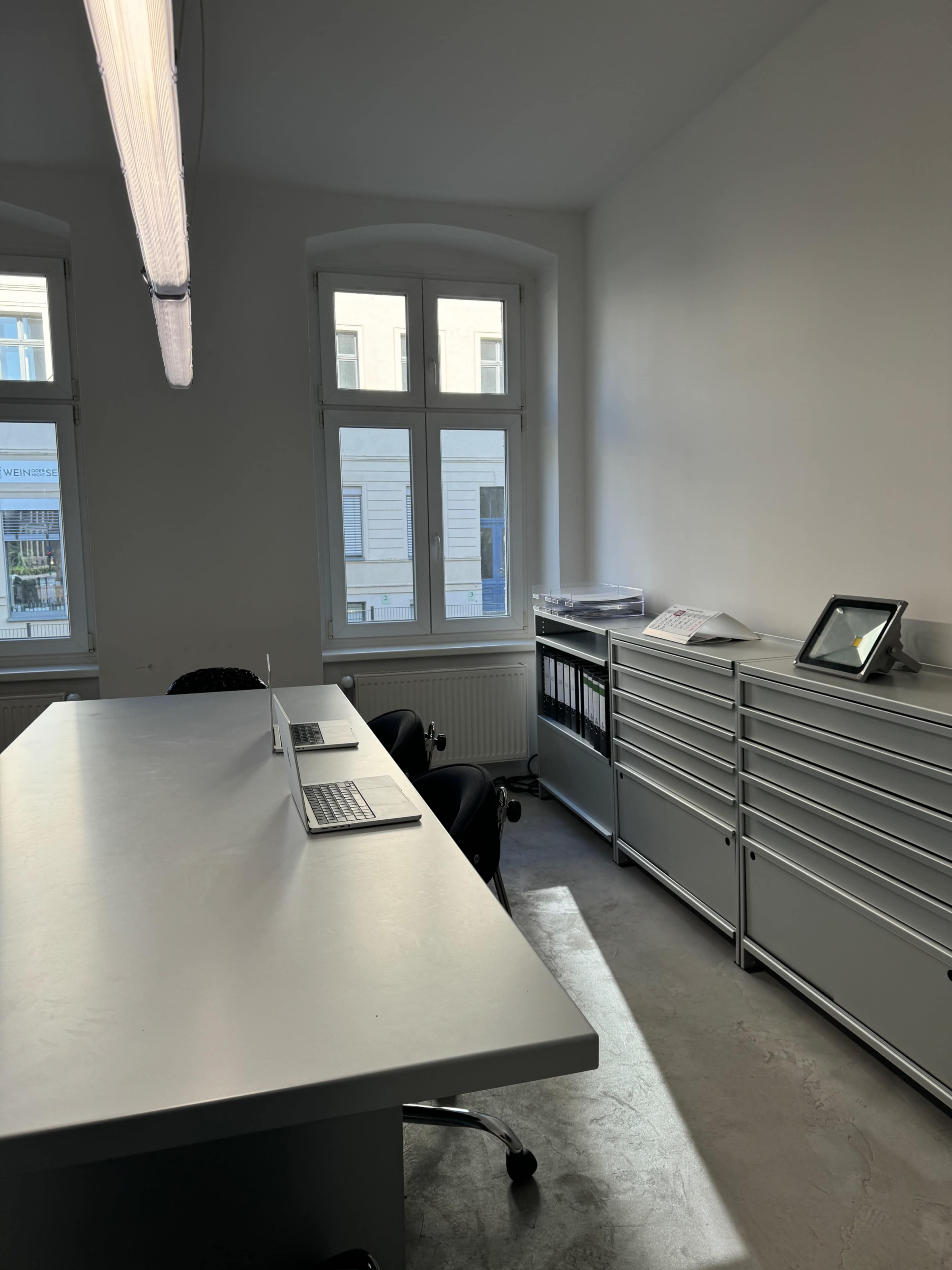 The image shows a modern office space with a long table, two laptops, ergonomic chairs, and metal storage cabinets beside large windows.