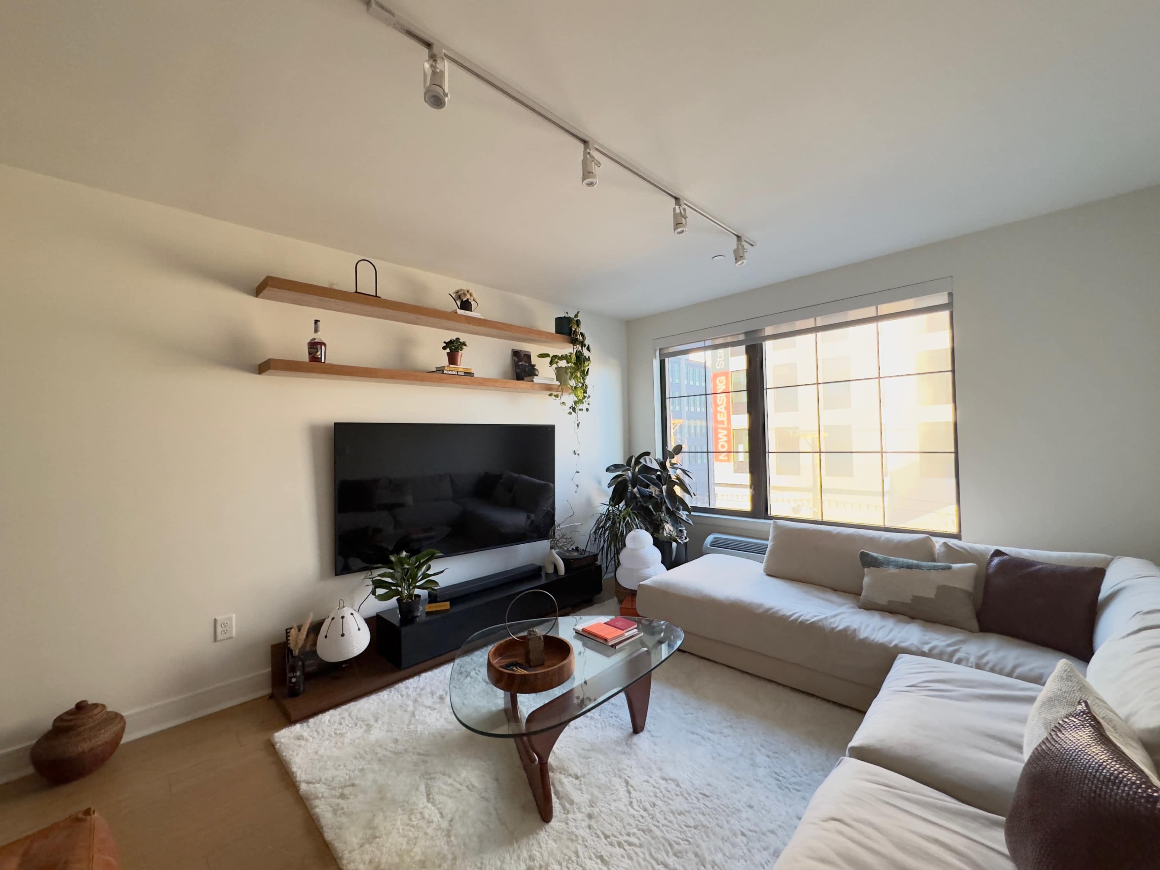 The living room features a large sectional sofa, a glass coffee table, and a wall-mounted television, with shelves displaying plants and decor.