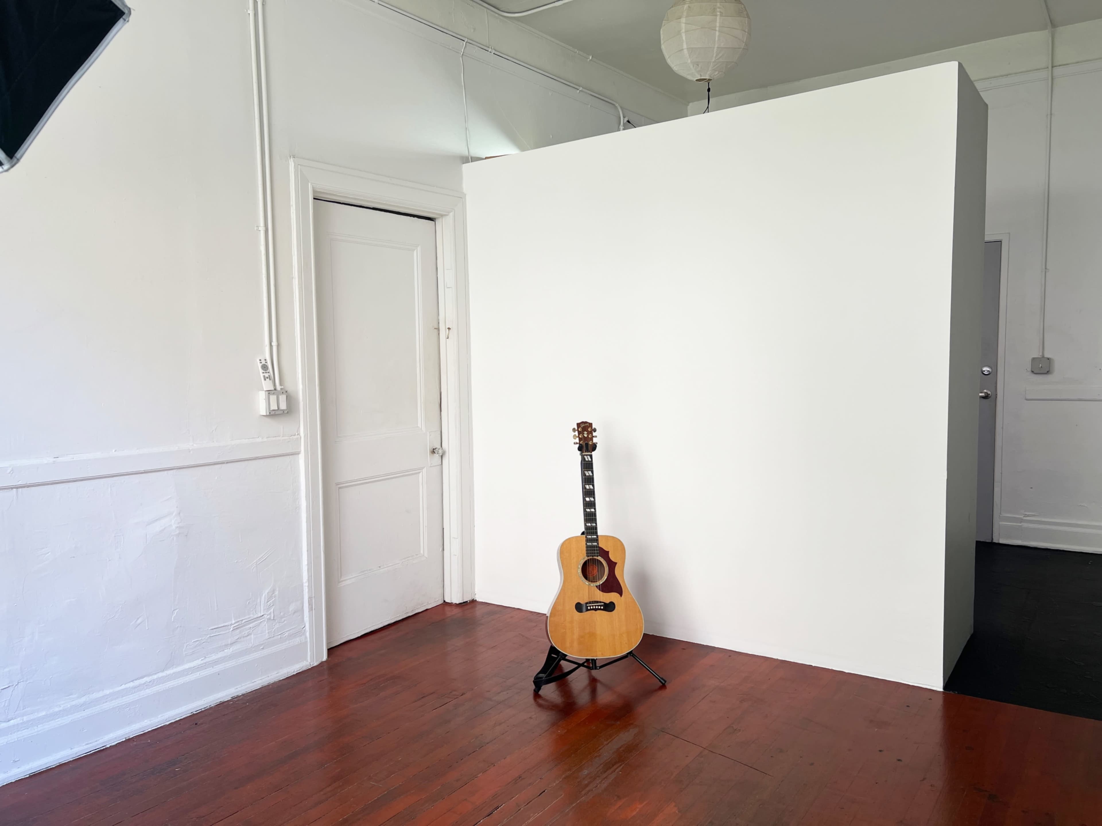 A wooden guitar stands on a stand in a sparsely furnished room with a white wall and a door.