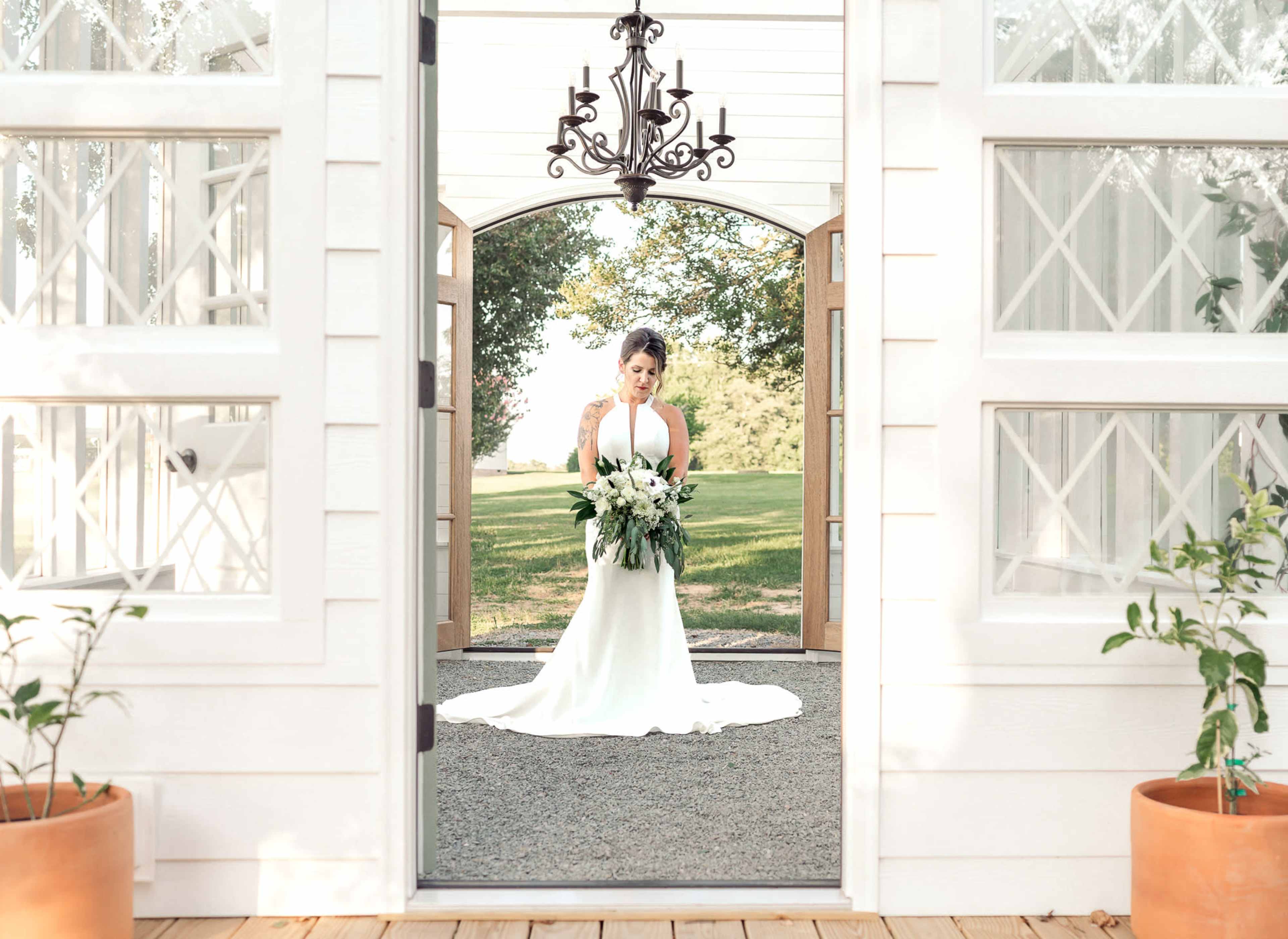 A bride in a white gown holds a bouquet while standing in a doorway with large wooden doors and a chandelier overhead.