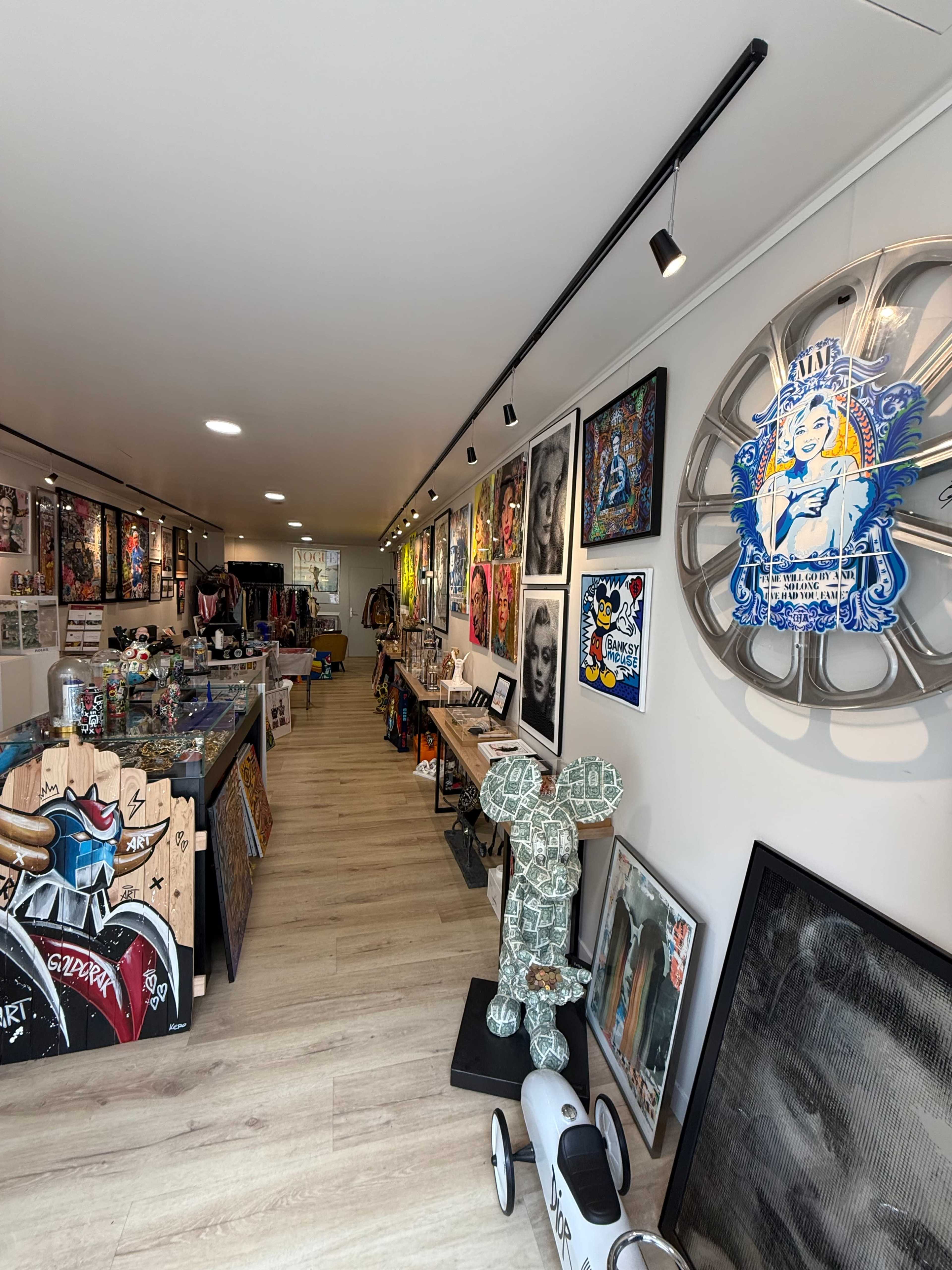 The image shows an art gallery with various artworks displayed on the walls and shelves, featuring colorful paintings and sculptures.