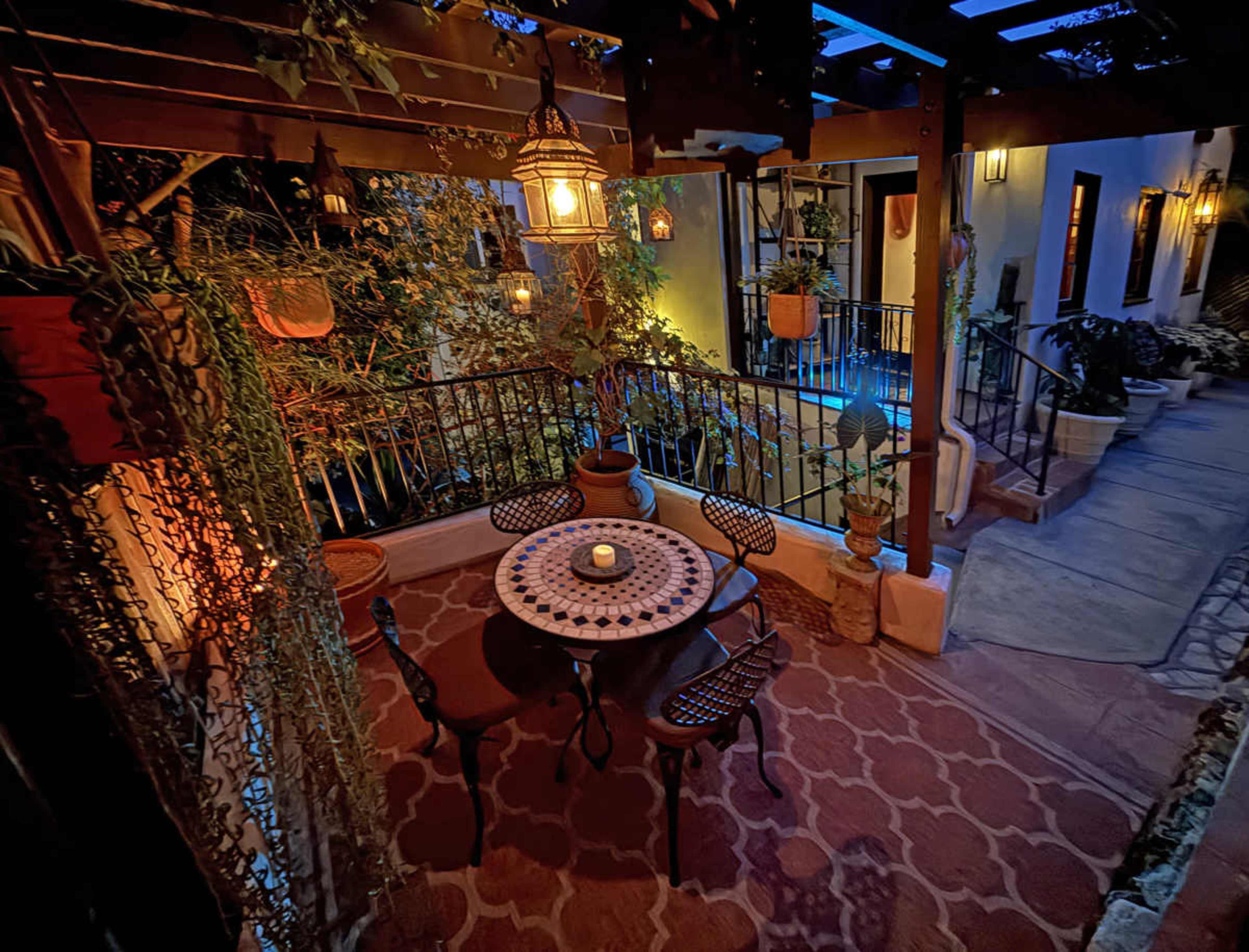 The image shows a warmly lit outdoor patio area with a round table surrounded by chairs, decorated with plants and lanterns under a wooden pergola.
