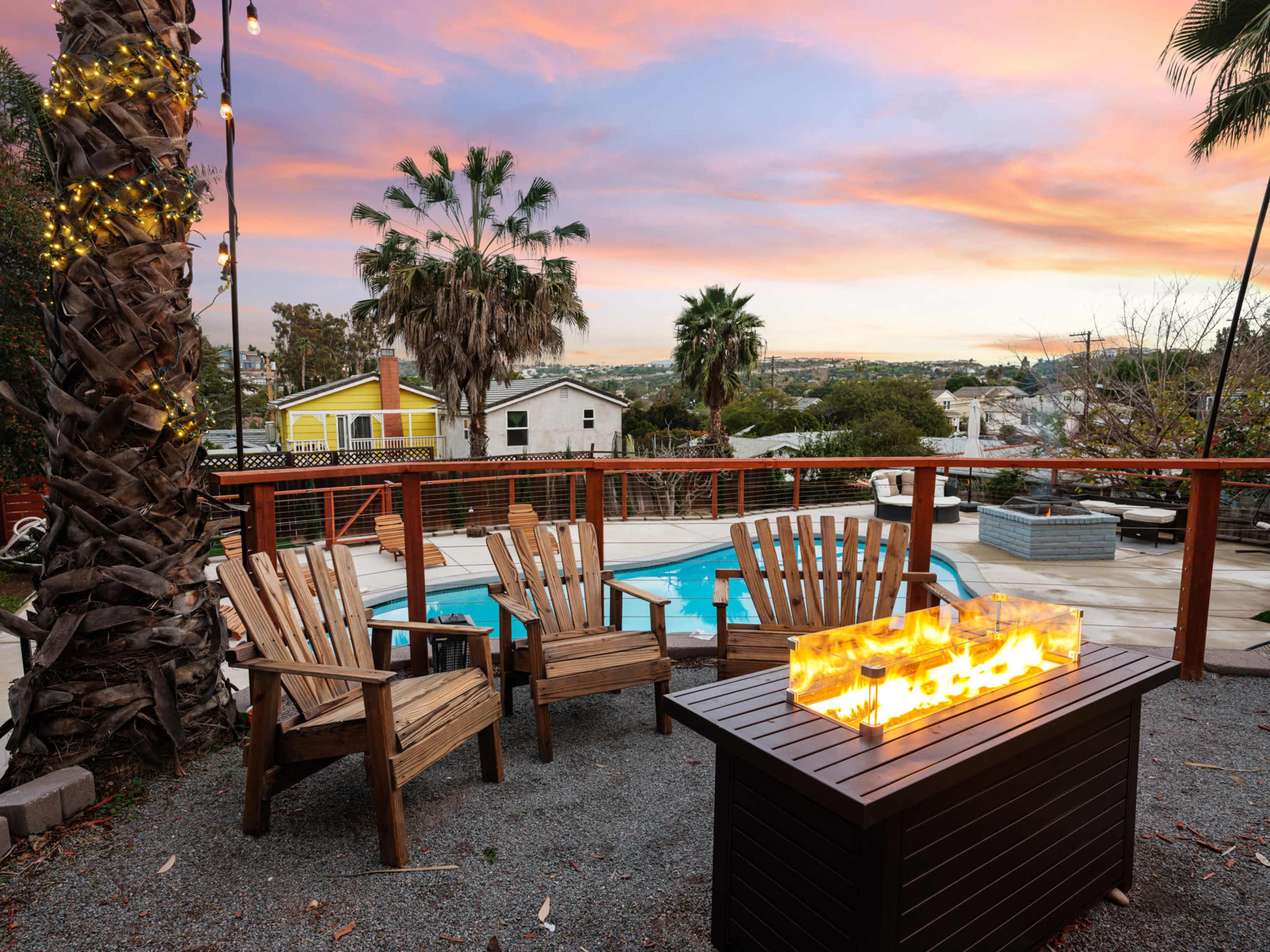 A small outdoor seating area with wooden chairs surrounds a fire pit, overlooking a pool and a sunset-lit landscape with palm trees.