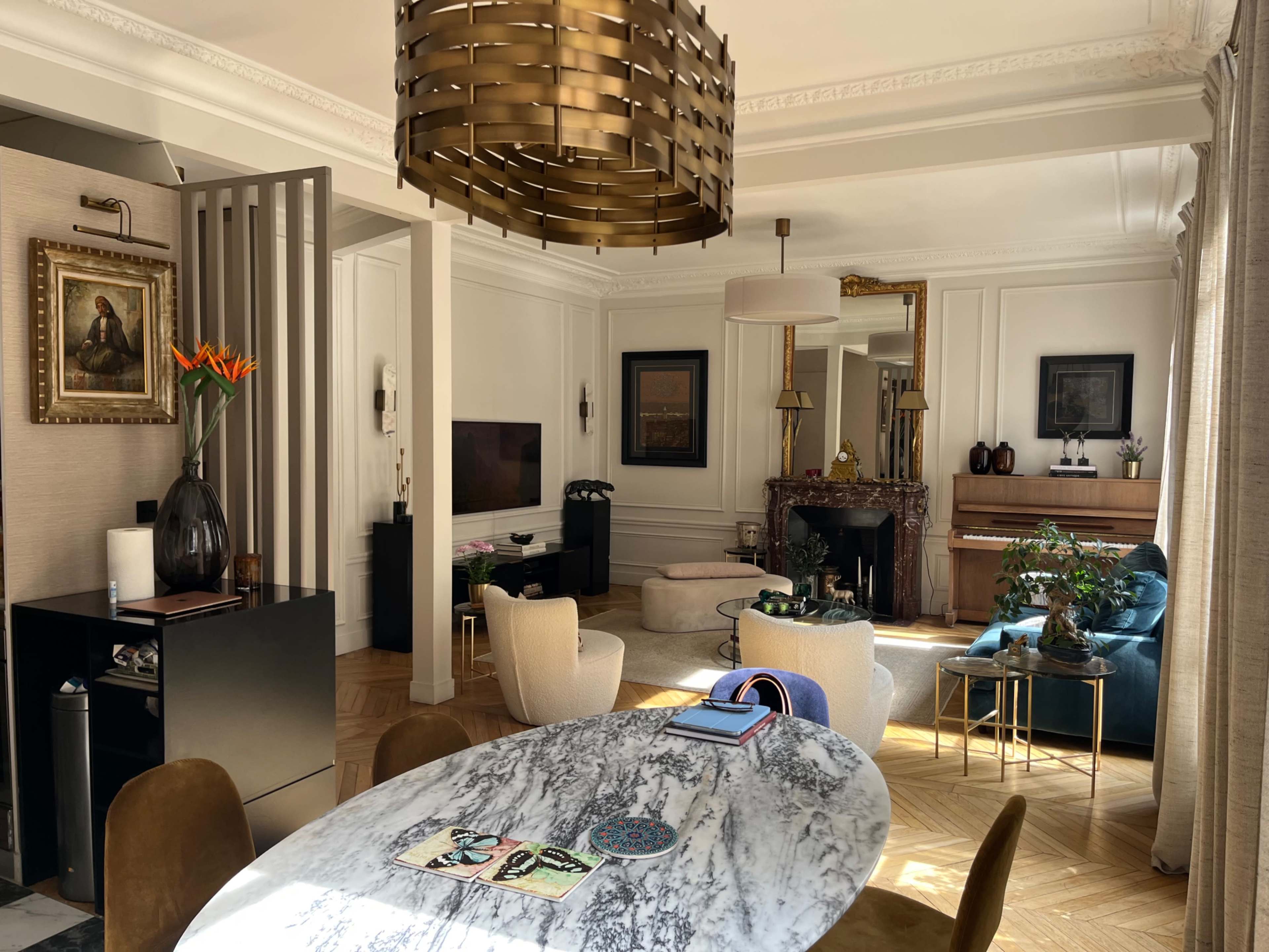 The image shows a well-furnished living room featuring a round marble table, various seating options, a television, and decorative items such as plants and artwork.
