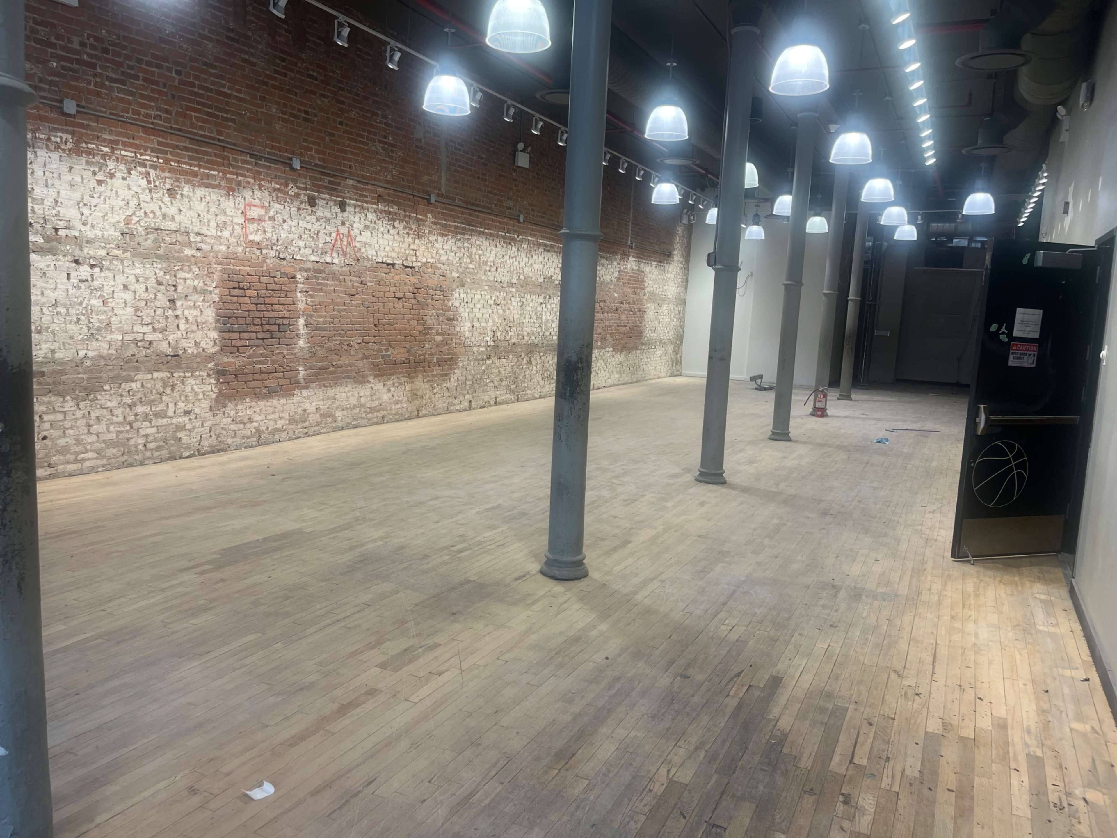 The image shows an empty, spacious room with exposed brick walls, wooden flooring, and several hanging light fixtures.