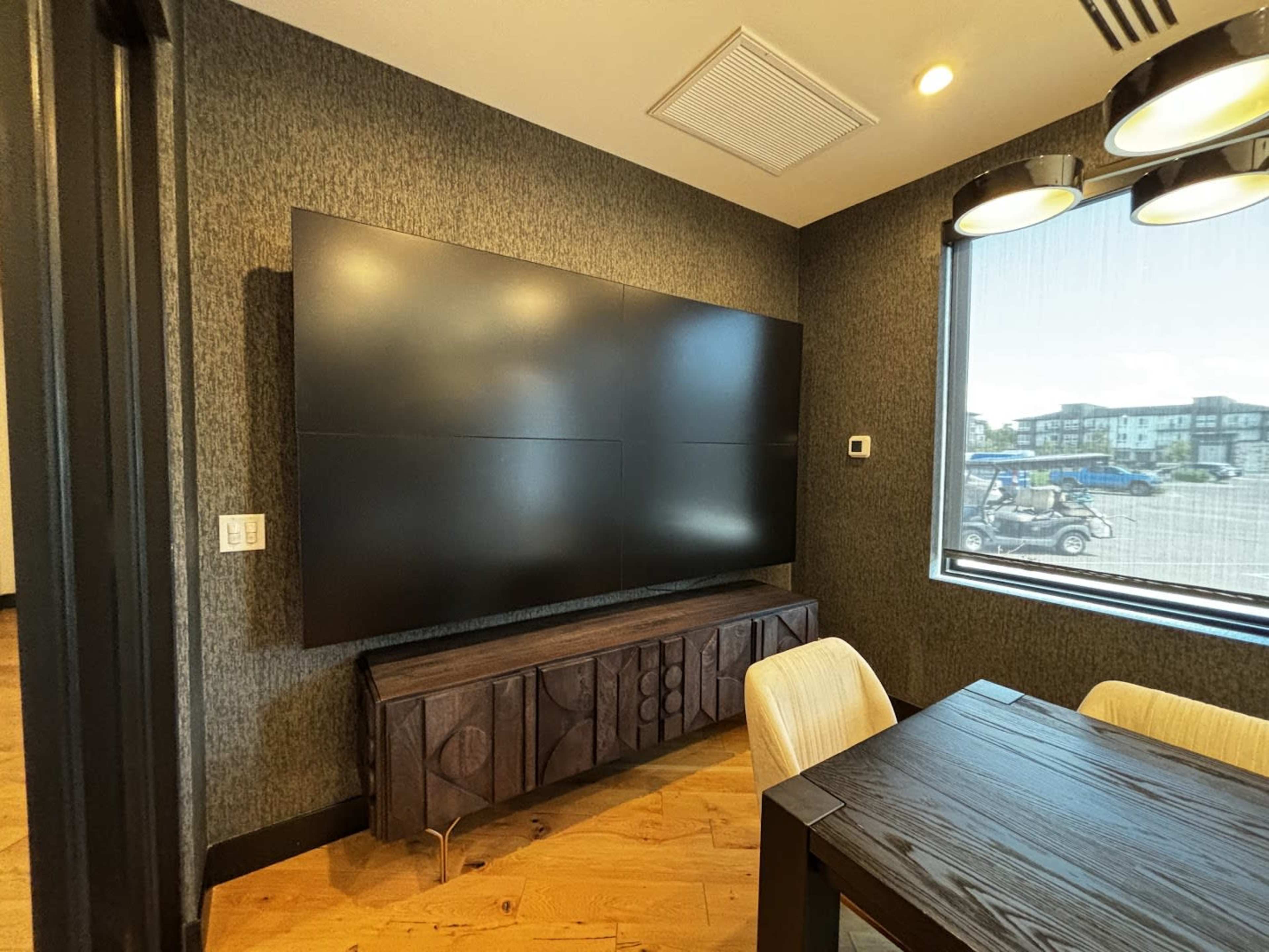 The image shows a conference room with a large wall-mounted screen above a wooden sideboard near a window.