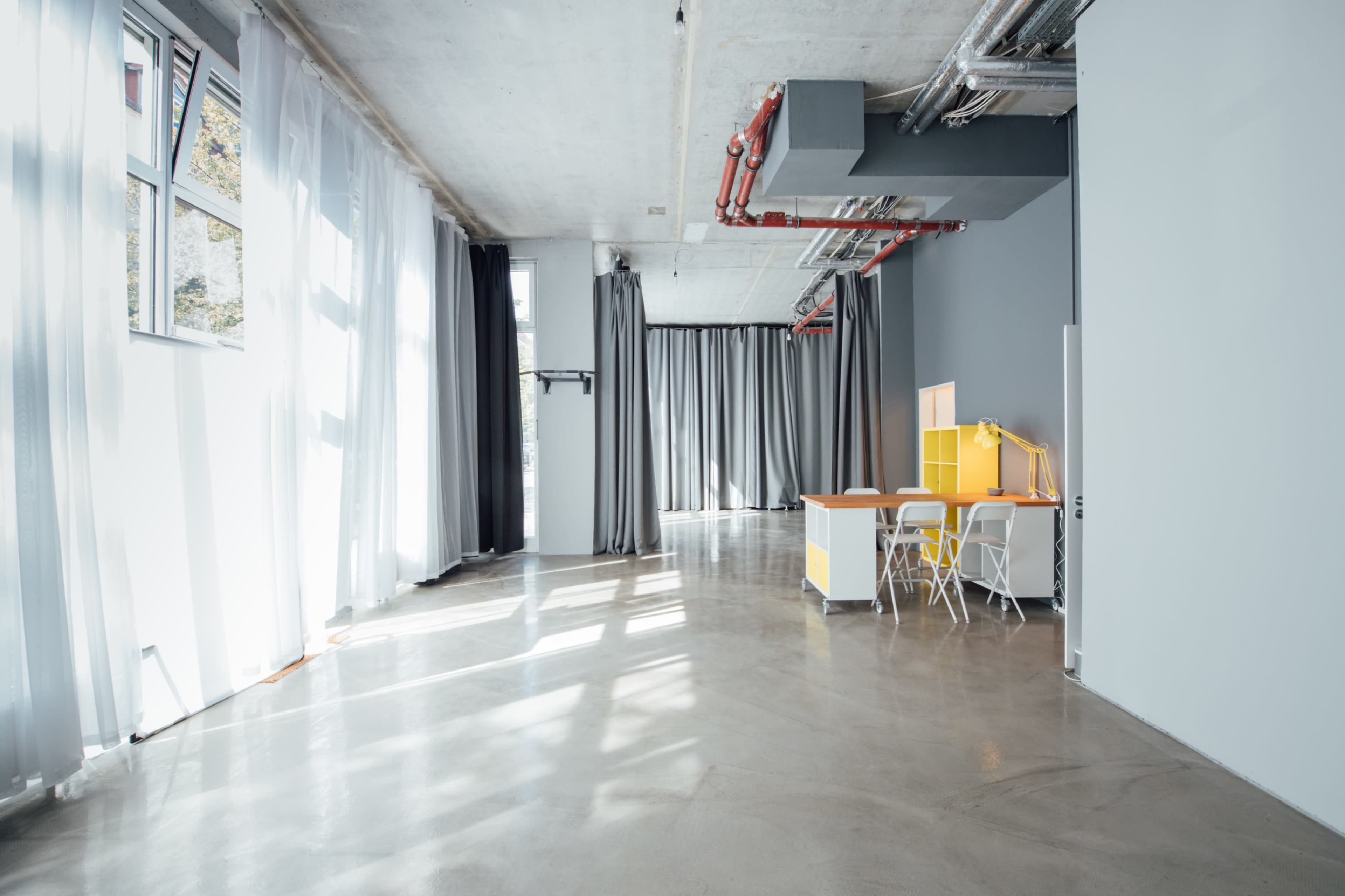 The image shows a spacious, modern interior with large windows, light curtains, and a minimalistic workspace featuring a yellow shelving unit and a table with chairs.