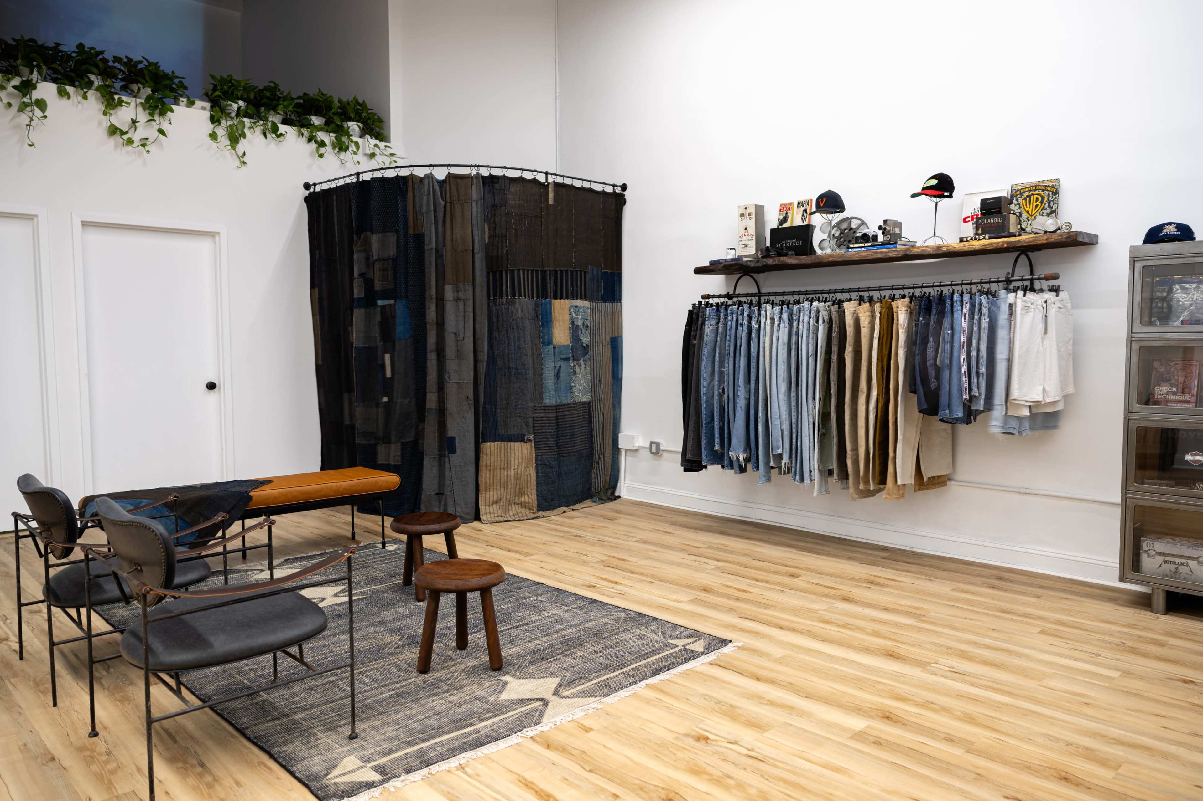 The image shows a minimalist clothing store interior featuring a display of denim items on a wall rack, a wooden table with two stools, and a patterned rug on the wooden floor.