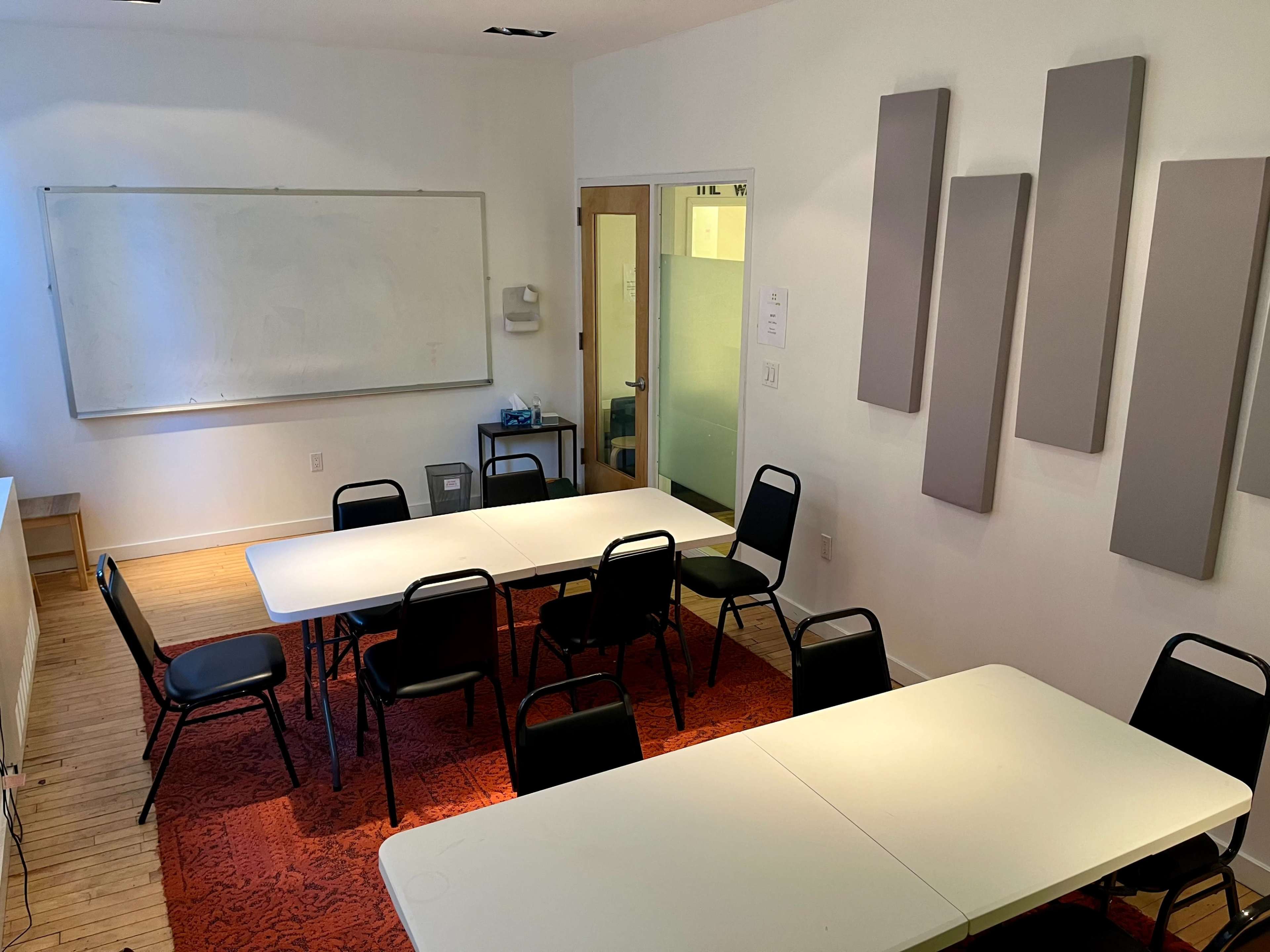 A classroom with two rectangular tables set up for seating, accompanied by several black chairs, a whiteboard on the wall, and sound-absorbing panels.