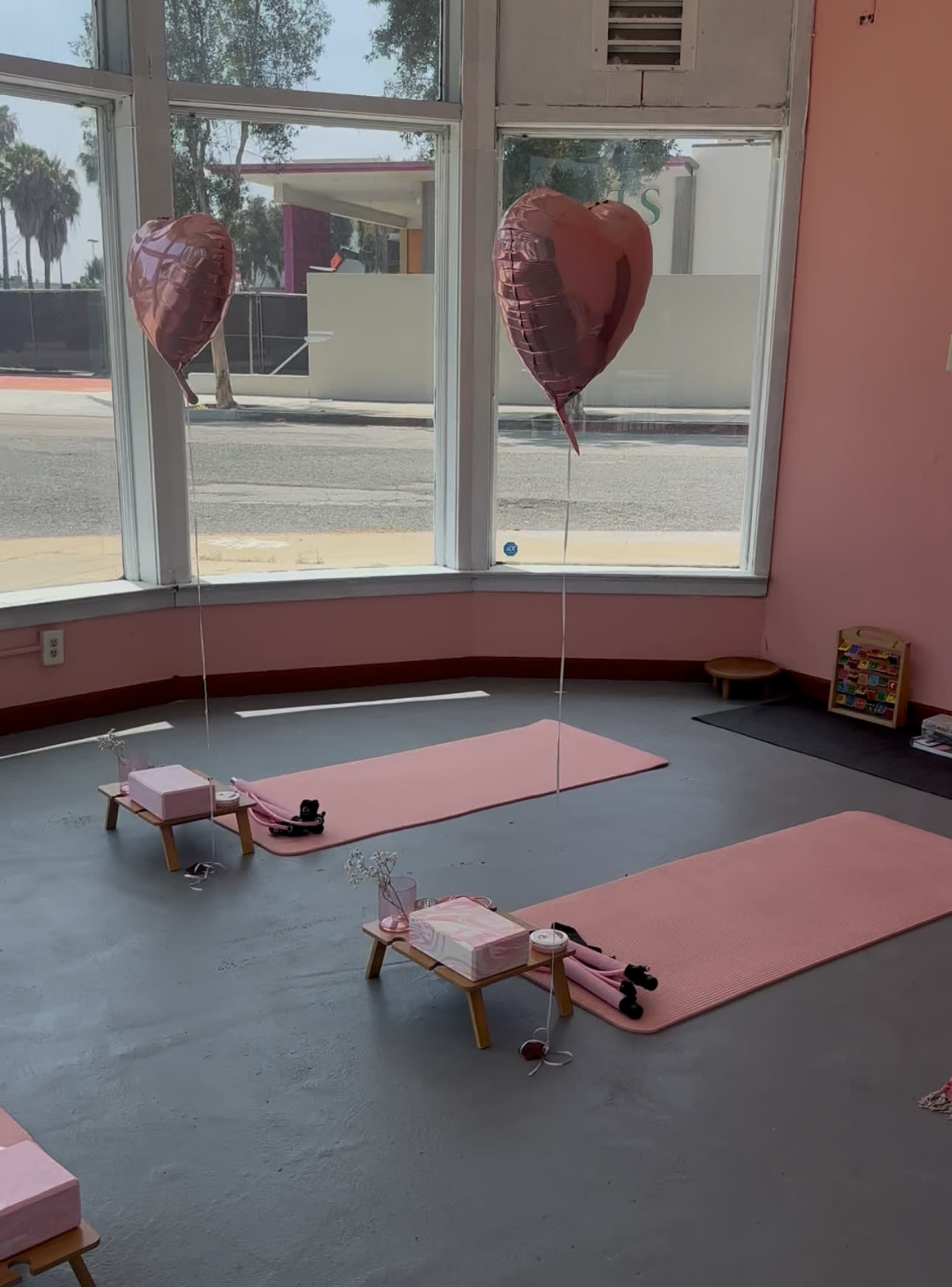 The image shows a pink-themed yoga studio with two pink mats, decorated with heart-shaped balloons and various yoga props.