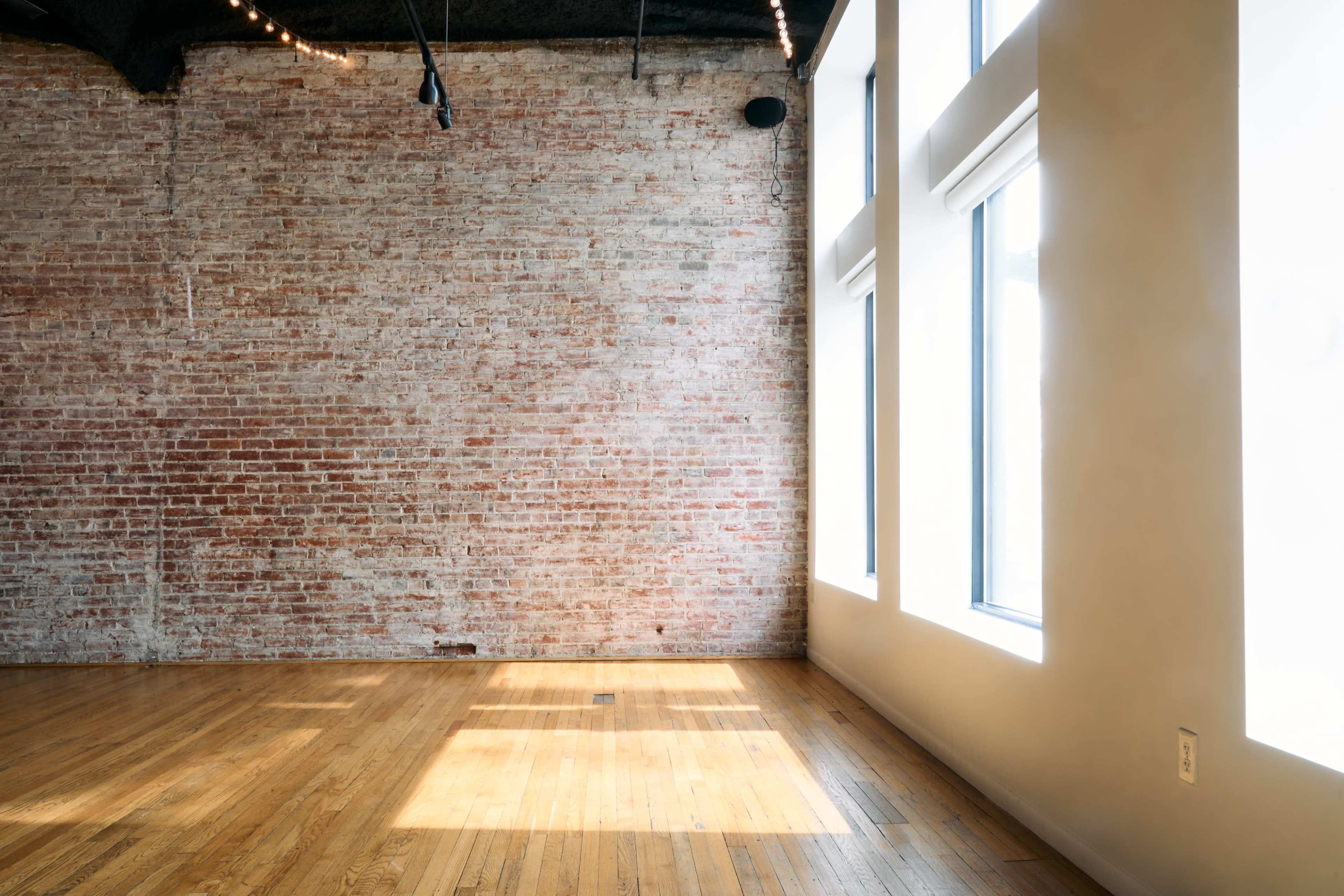 The image shows a spacious interior with exposed brick walls and large windows allowing natural light to enter, alongside a hardwood floor.
