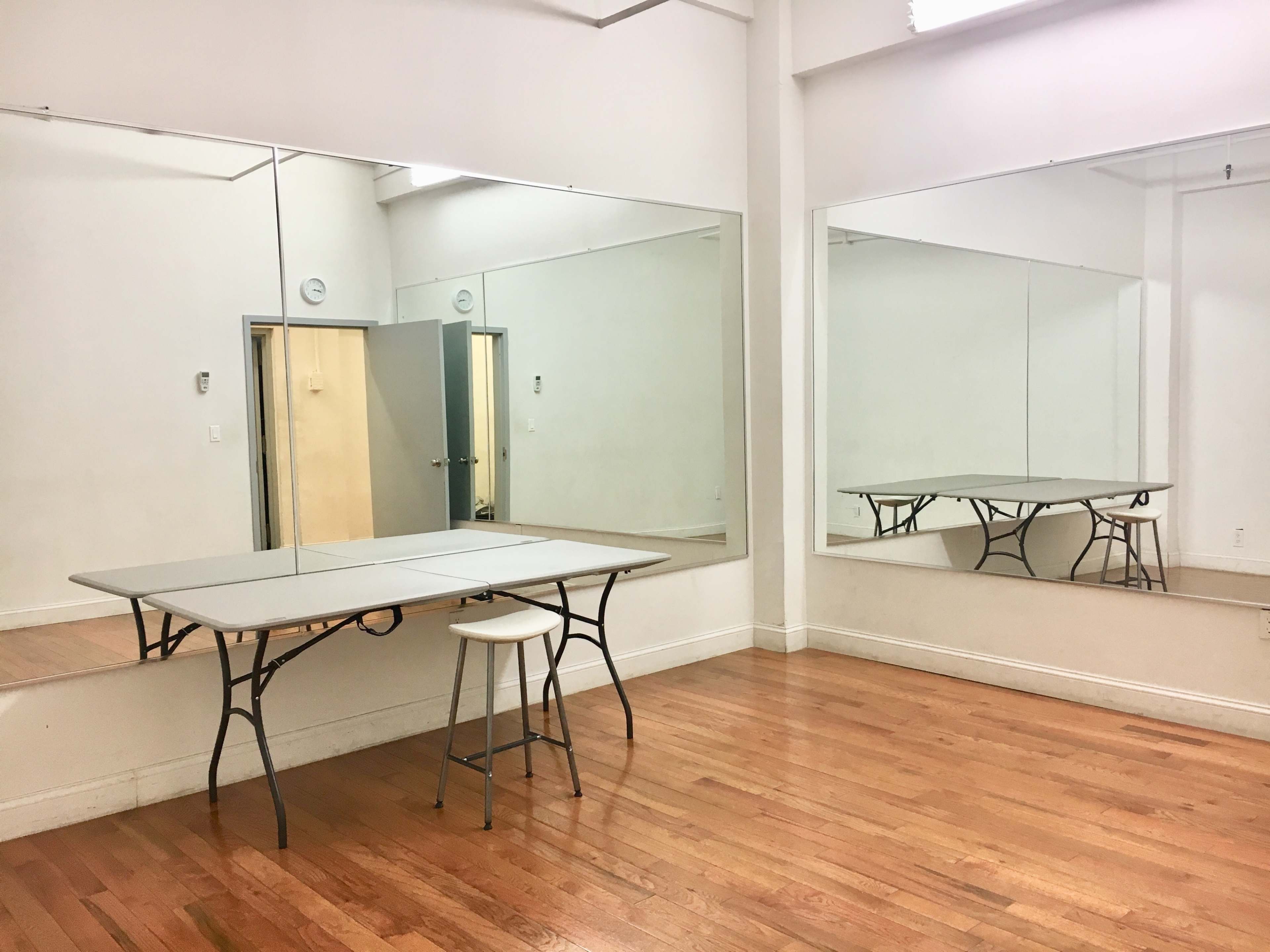A sparsely furnished room with a folding table and a stool, featuring large mirrors on two walls and hardwood flooring.