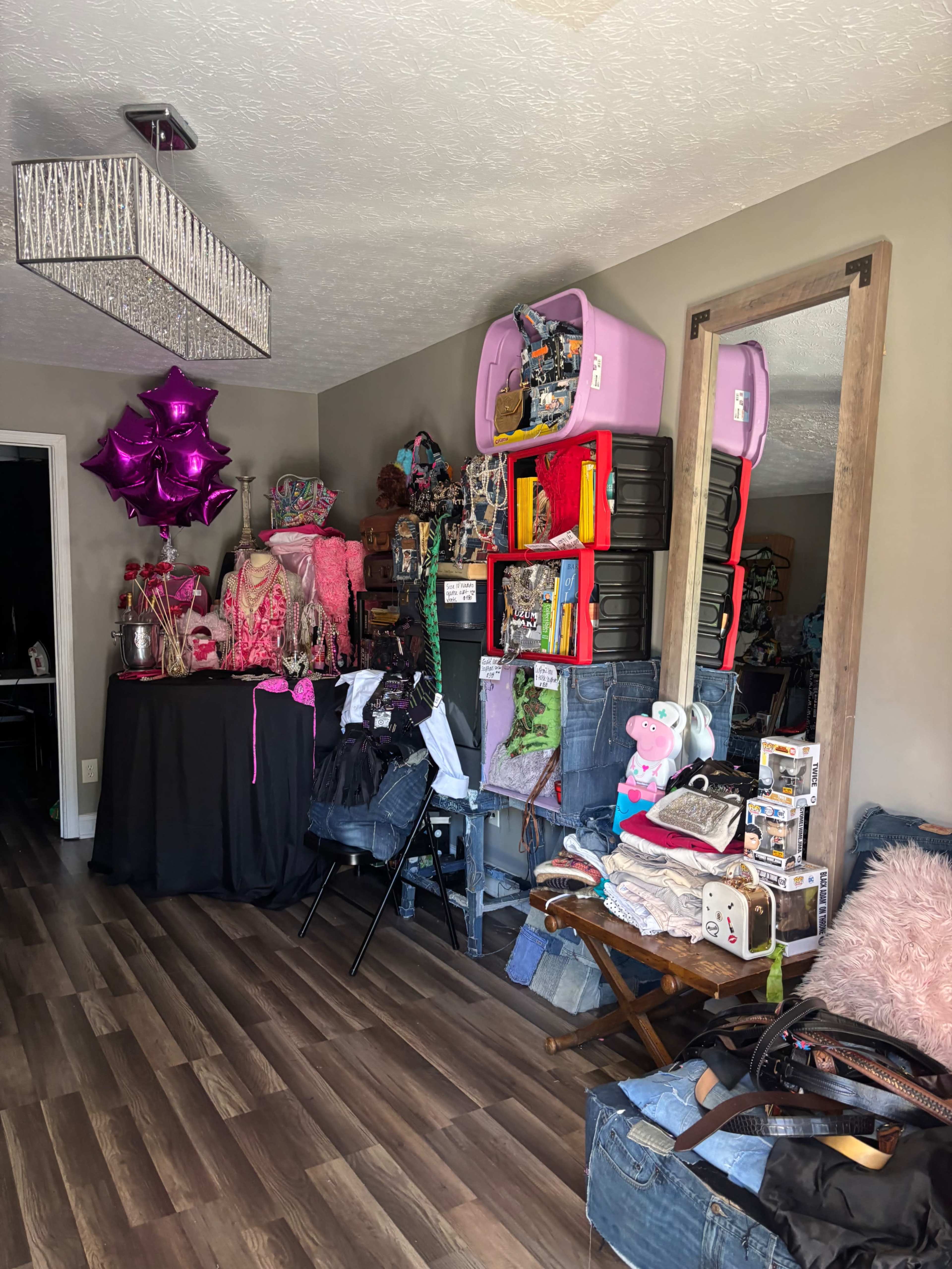 The image shows a cluttered living space with various items, including stacked boxes, a table with decorations, and a large mirror against the wall.