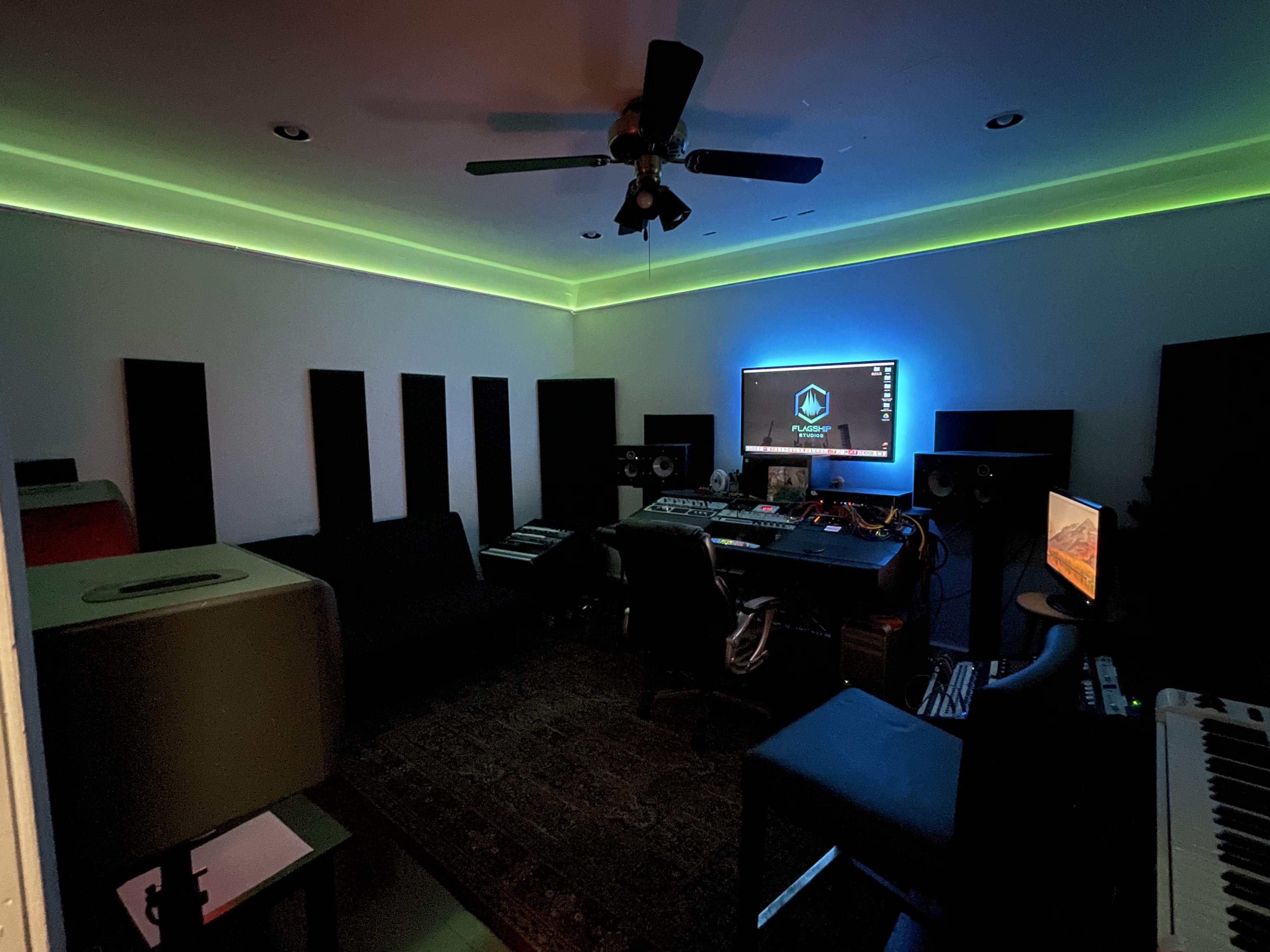 A music production studio features a computer setup with multiple monitors, synthesizers, and soundproof panels, illuminated by colored LED lights along the ceiling.