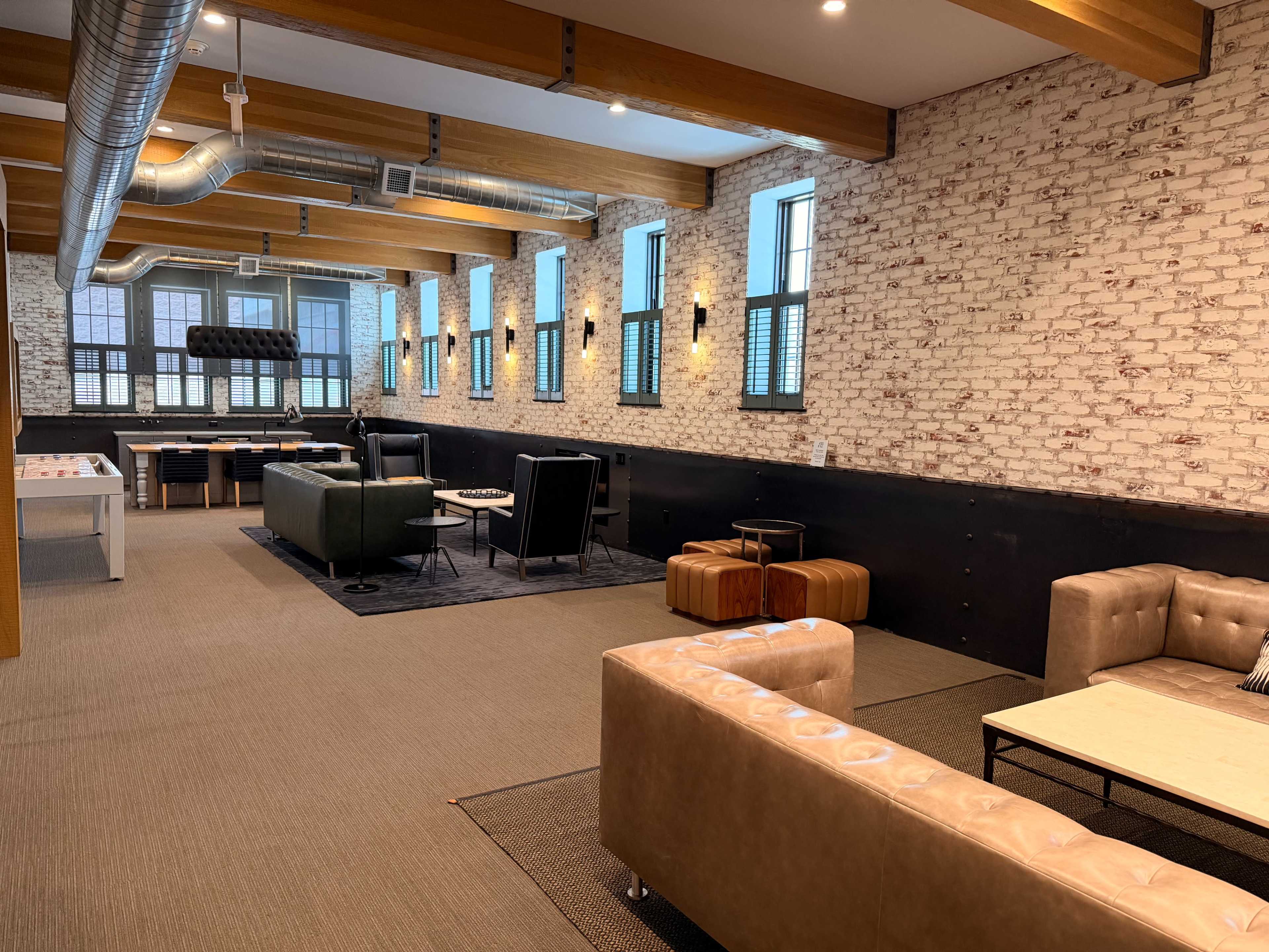 The image shows a large, industrial-style room with exposed brick walls, multiple seating areas, and large windows allowing natural light to enter.
