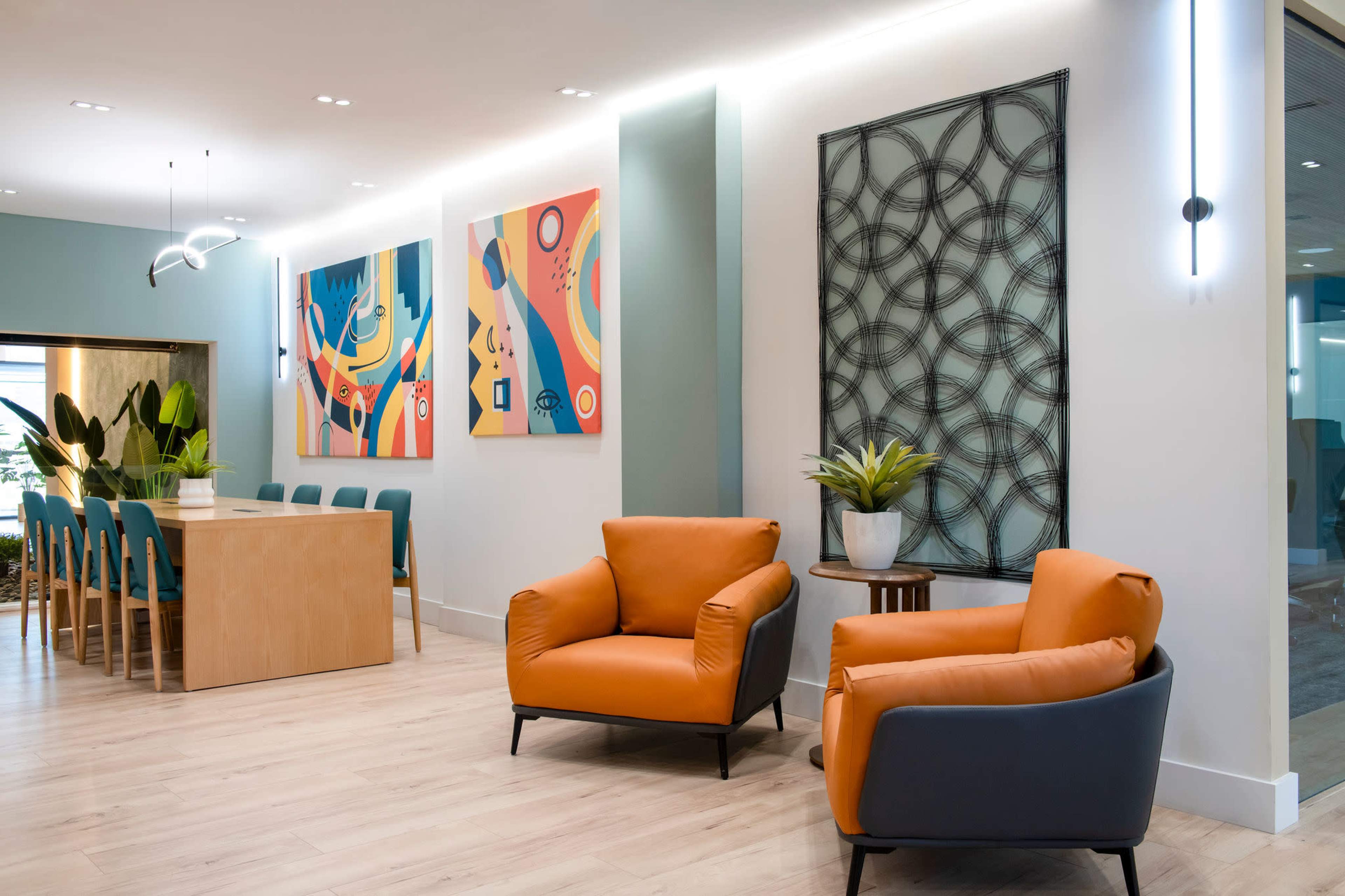 The image shows a modern interior space featuring a dining area with a large wooden table, surrounded by blue chairs, and two orange armchairs in a lounge area adorned with colorful wall art.
