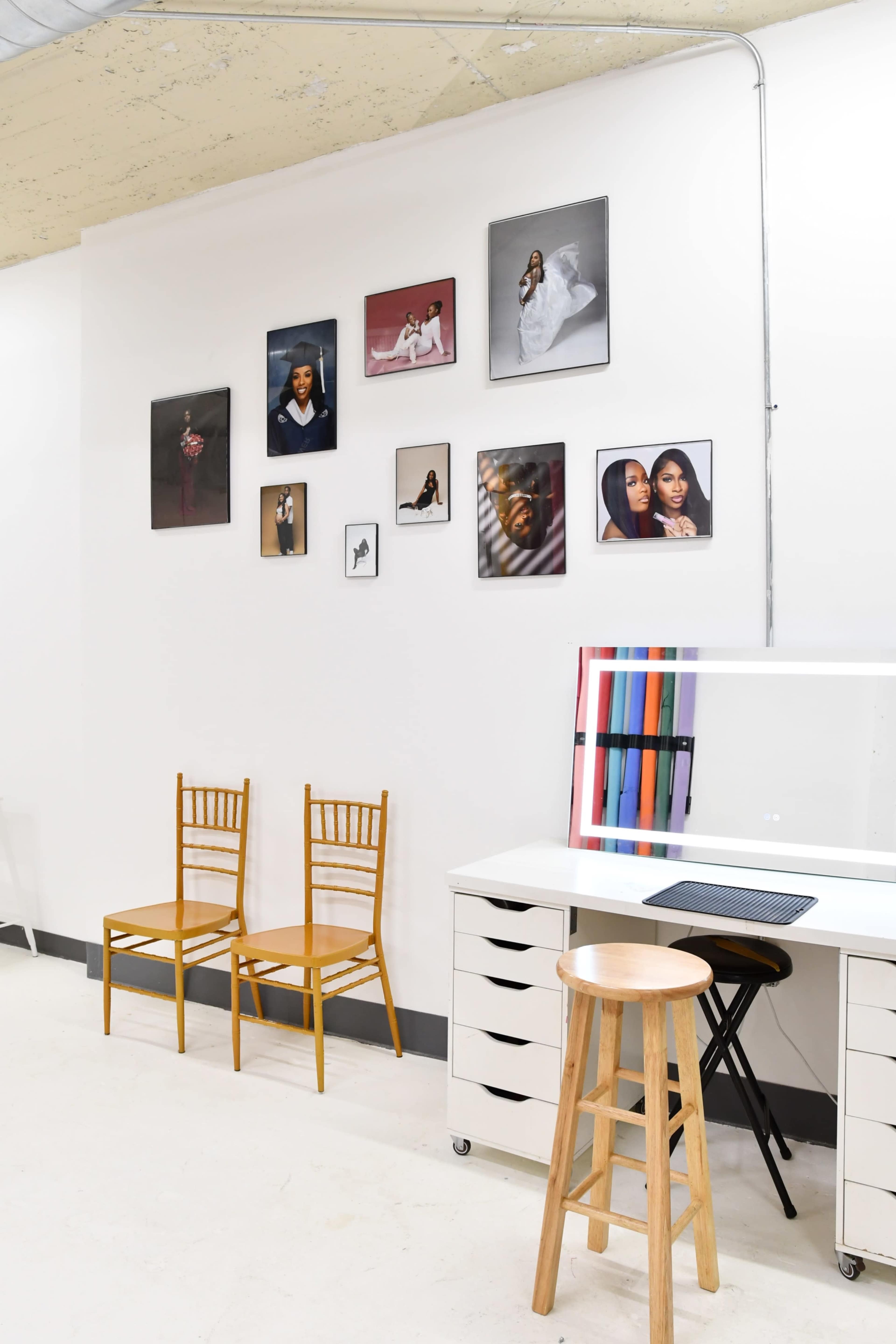 The scene features a minimalist studio space with two wooden chairs, a desk with a stool, and a gallery wall displaying various framed photographs.