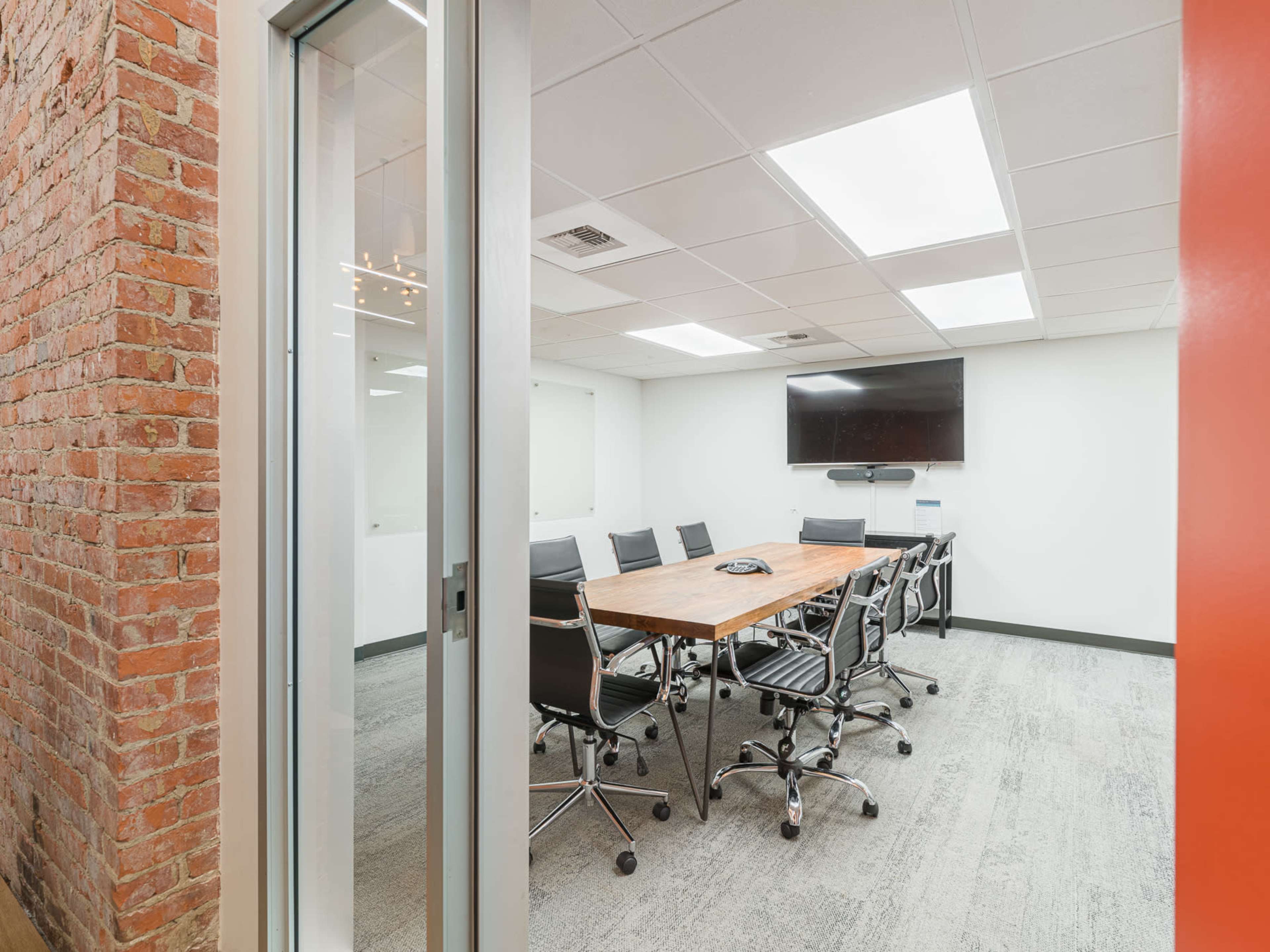 The image shows a modern conference room featuring a large wooden table surrounded by black office chairs, a wall-mounted television, and exposed brick on one side.