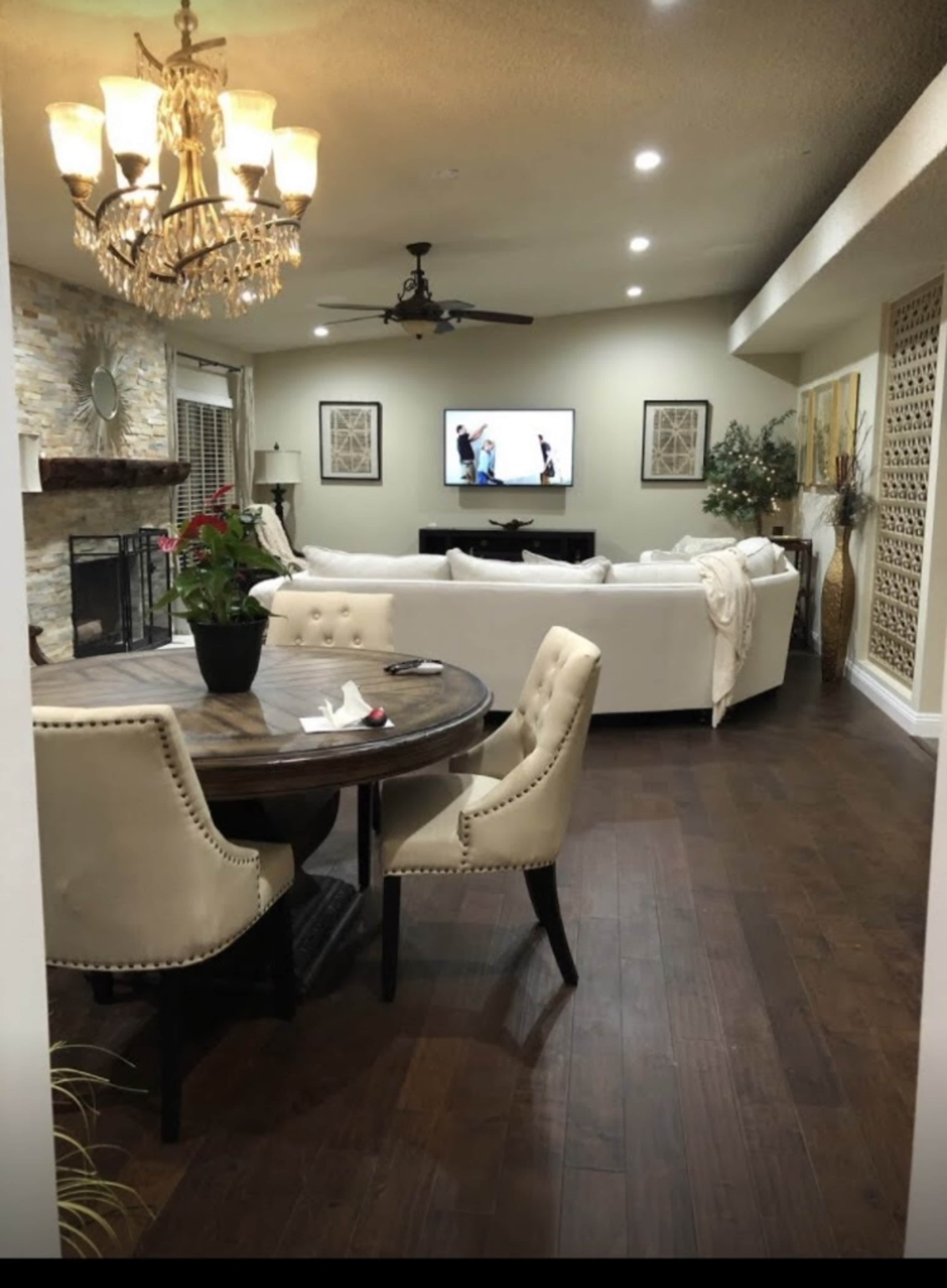 The image shows a living room with a chandelier, a TV mounted on the wall, a large white sofa, and a round dining table with upholstered chairs.