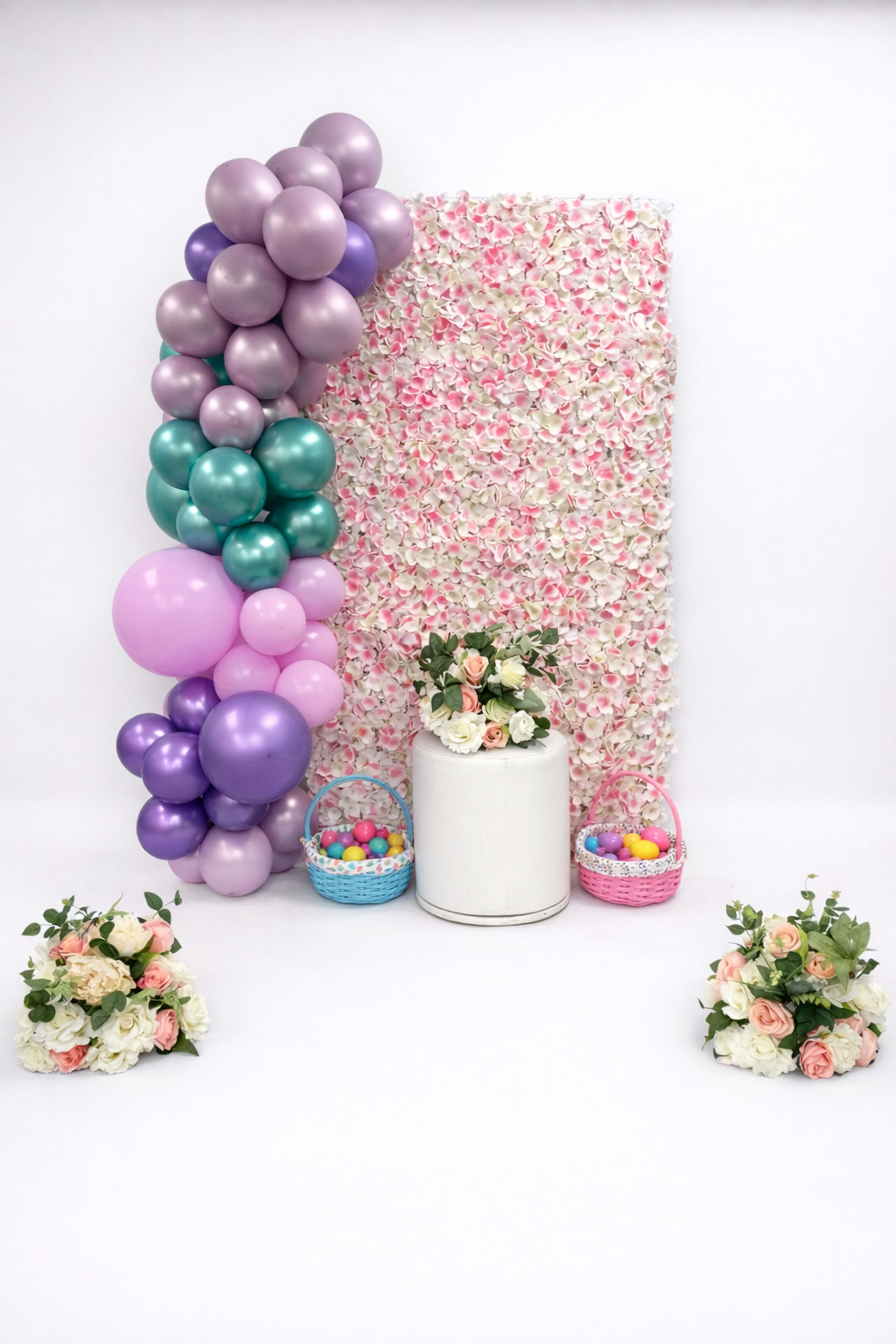 The image features a festive backdrop of floral decorations, accompanied by colorful balloons, a white pedestal with flowers, and two baskets filled with brightly colored eggs.