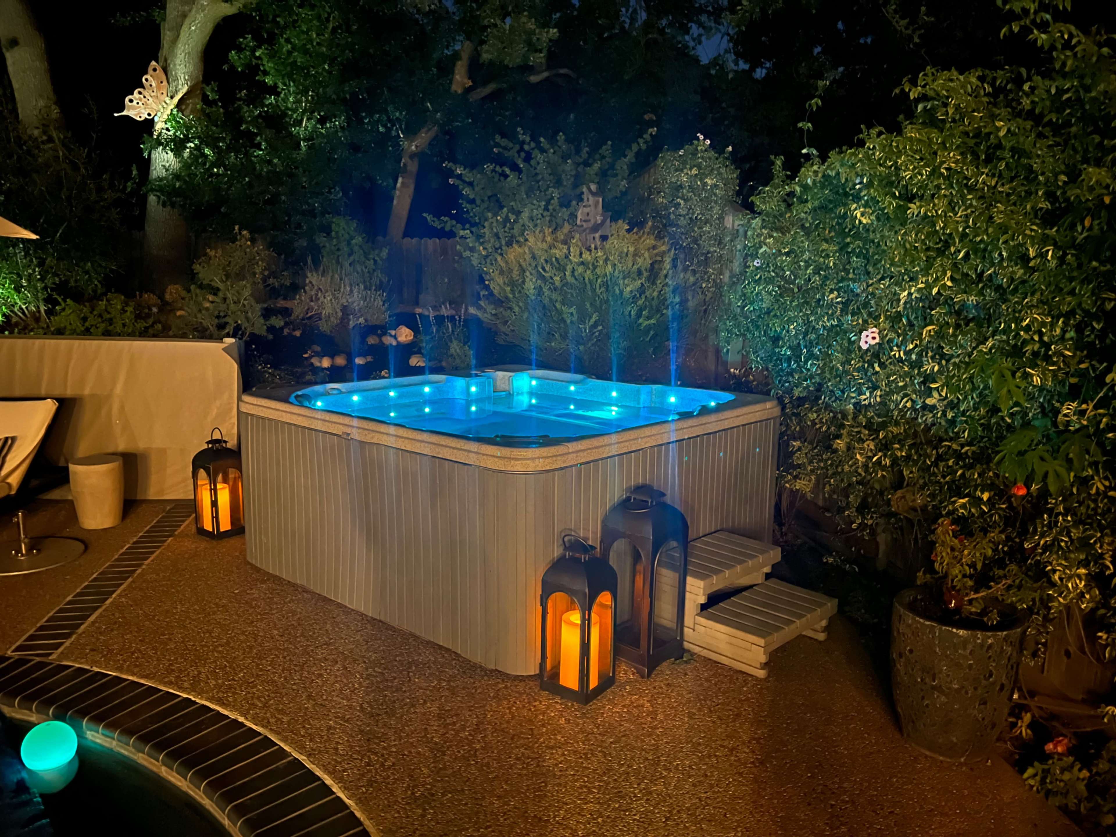 A hot tub with blue lights is positioned in a garden surrounded by plants and lanterns at night.