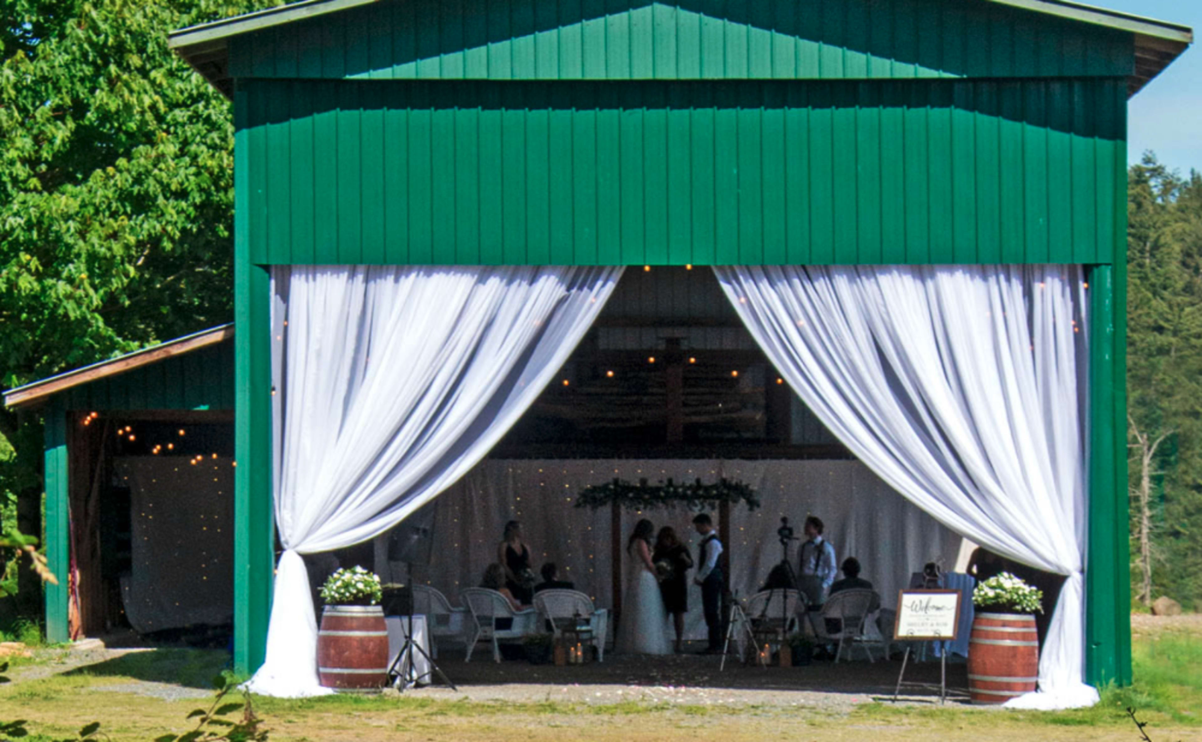 A wedding ceremony takes place under a large, draped awning in a rustic green barn.