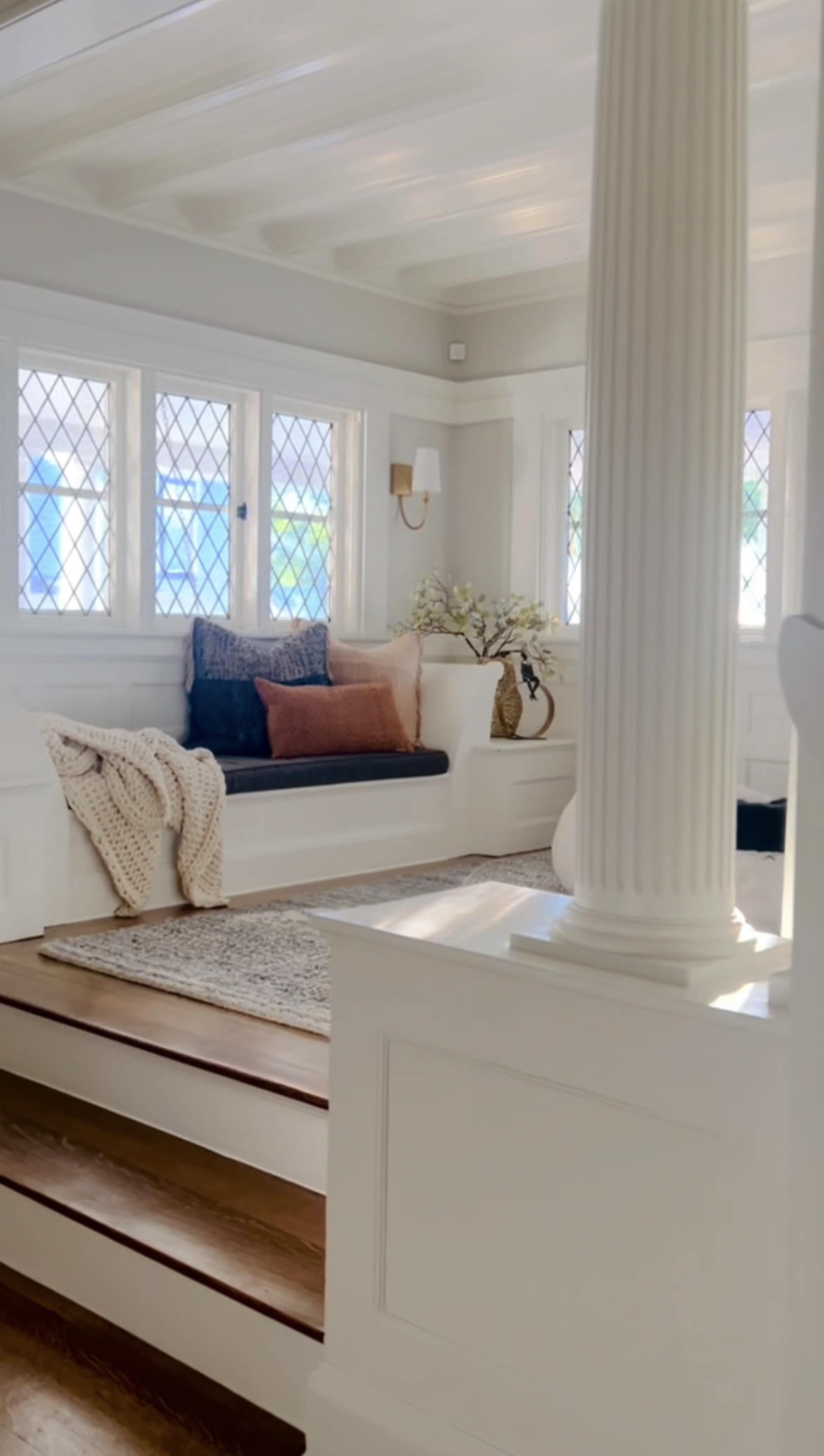 The image shows a well-lit living space featuring a white frame, a dark cushioned bench with pillows, and decorative elements like a vase and a cozy blanket.