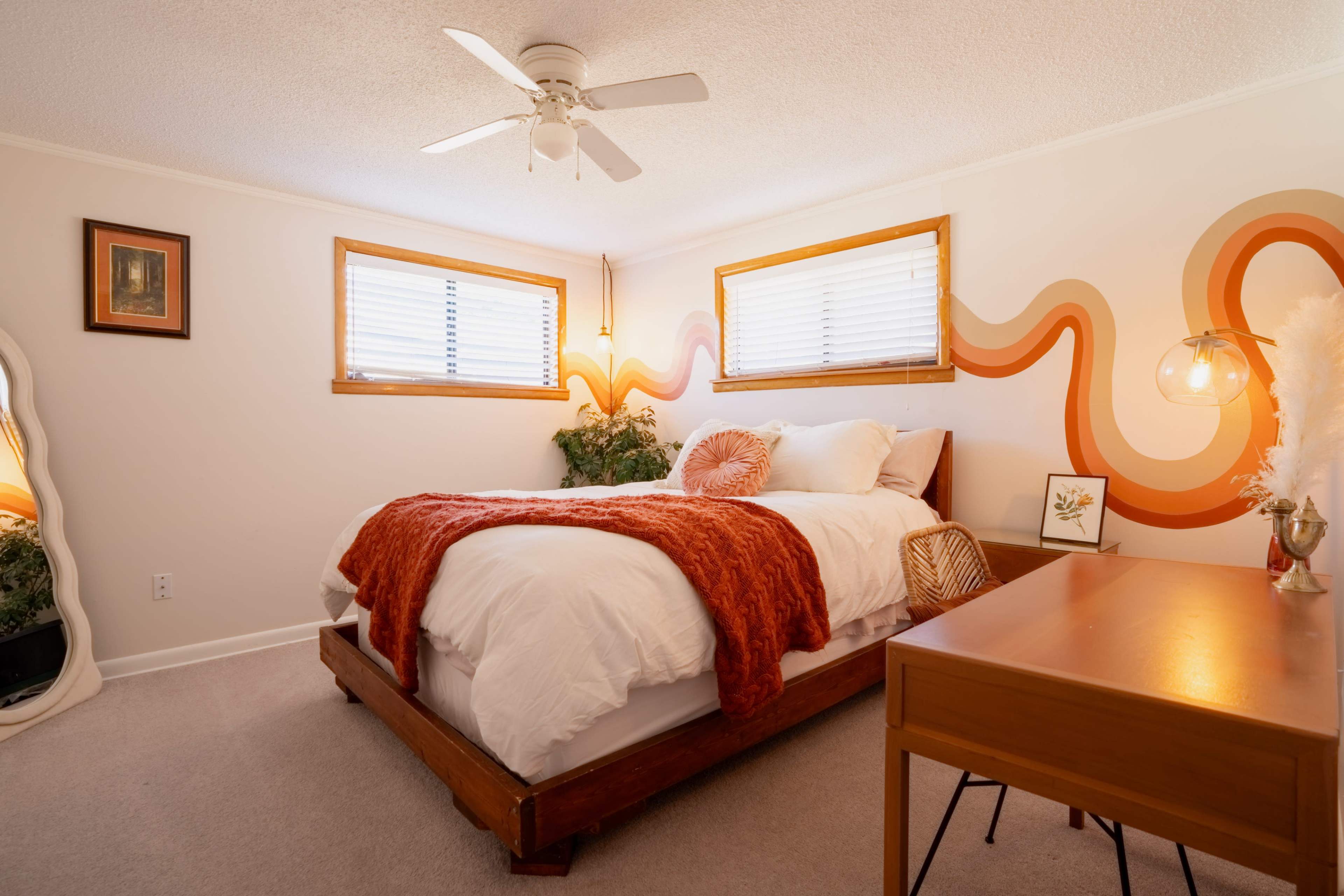 The image depicts a cozy bedroom featuring a wooden bed with white bedding, an orange throw blanket, a desk, and windows adorned with wooden blinds.