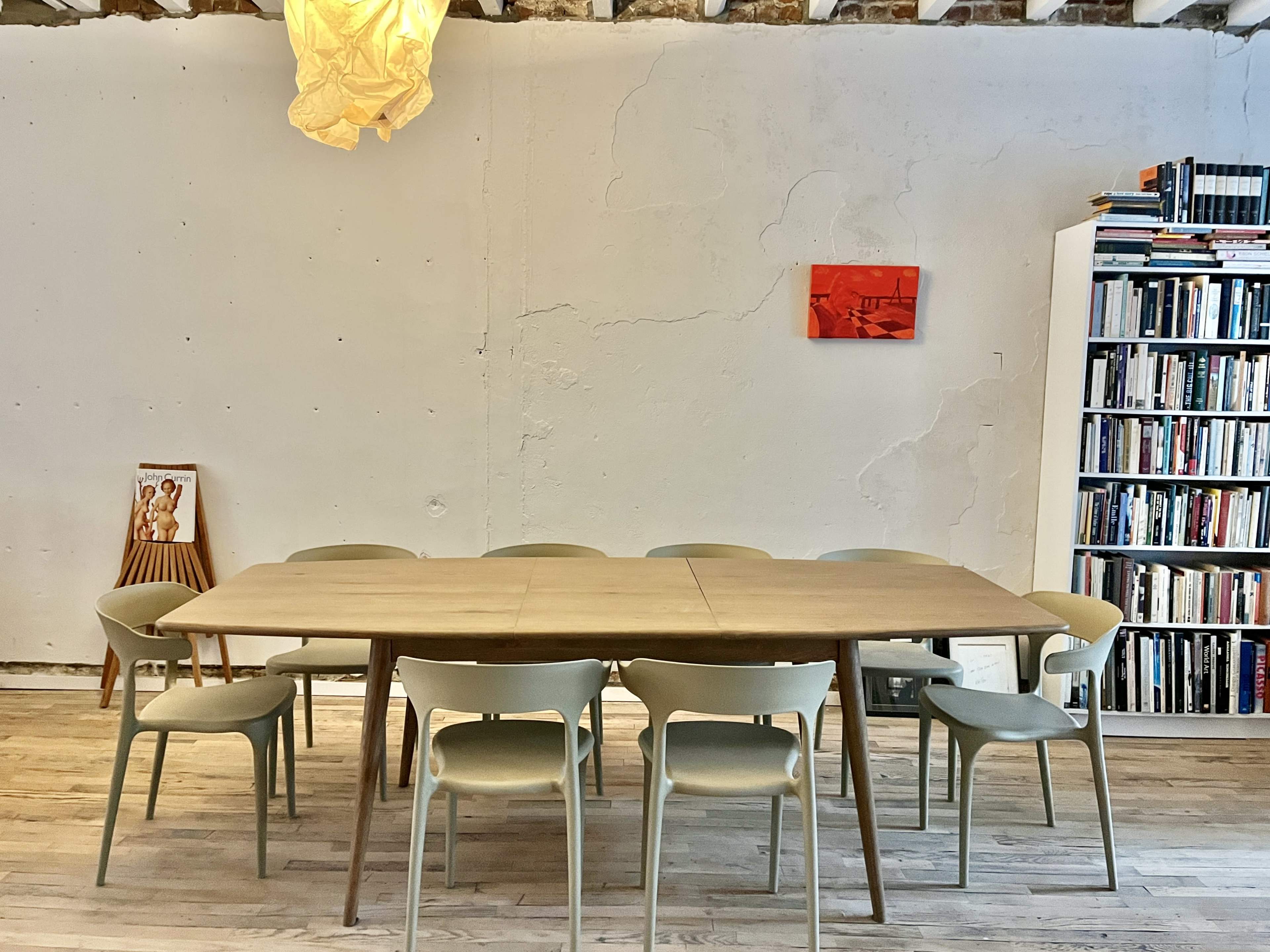A wooden dining table surrounded by eight light-colored chairs is positioned in a room with a textured wall, a pendant light above, and a bookshelf filled with books in the background.