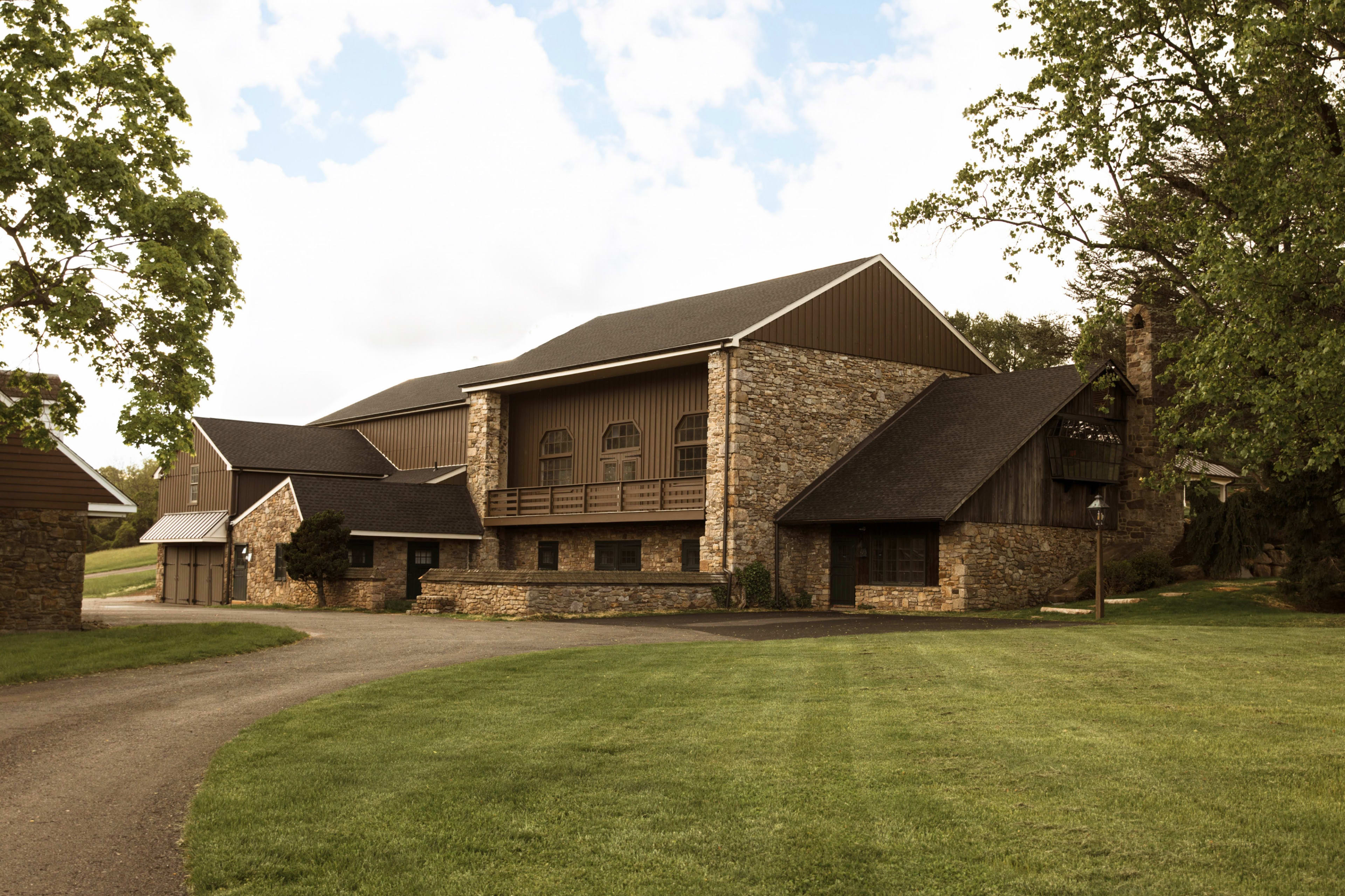 The image shows a large stone and wood building surrounded by a grassy area and trees, with a winding driveway leading to its entrance.