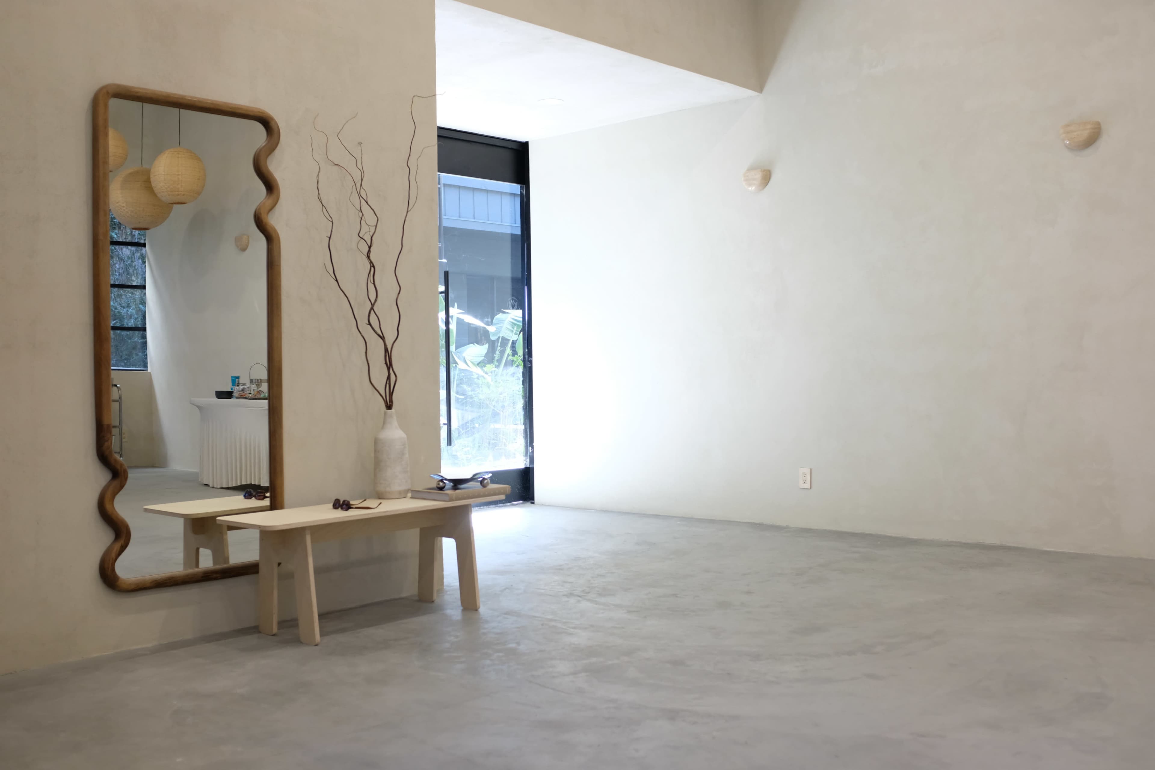 The image shows a minimalist interior space with a large, wavy-framed mirror, a simple wooden bench, and decorative elements like a vase and slender branches.