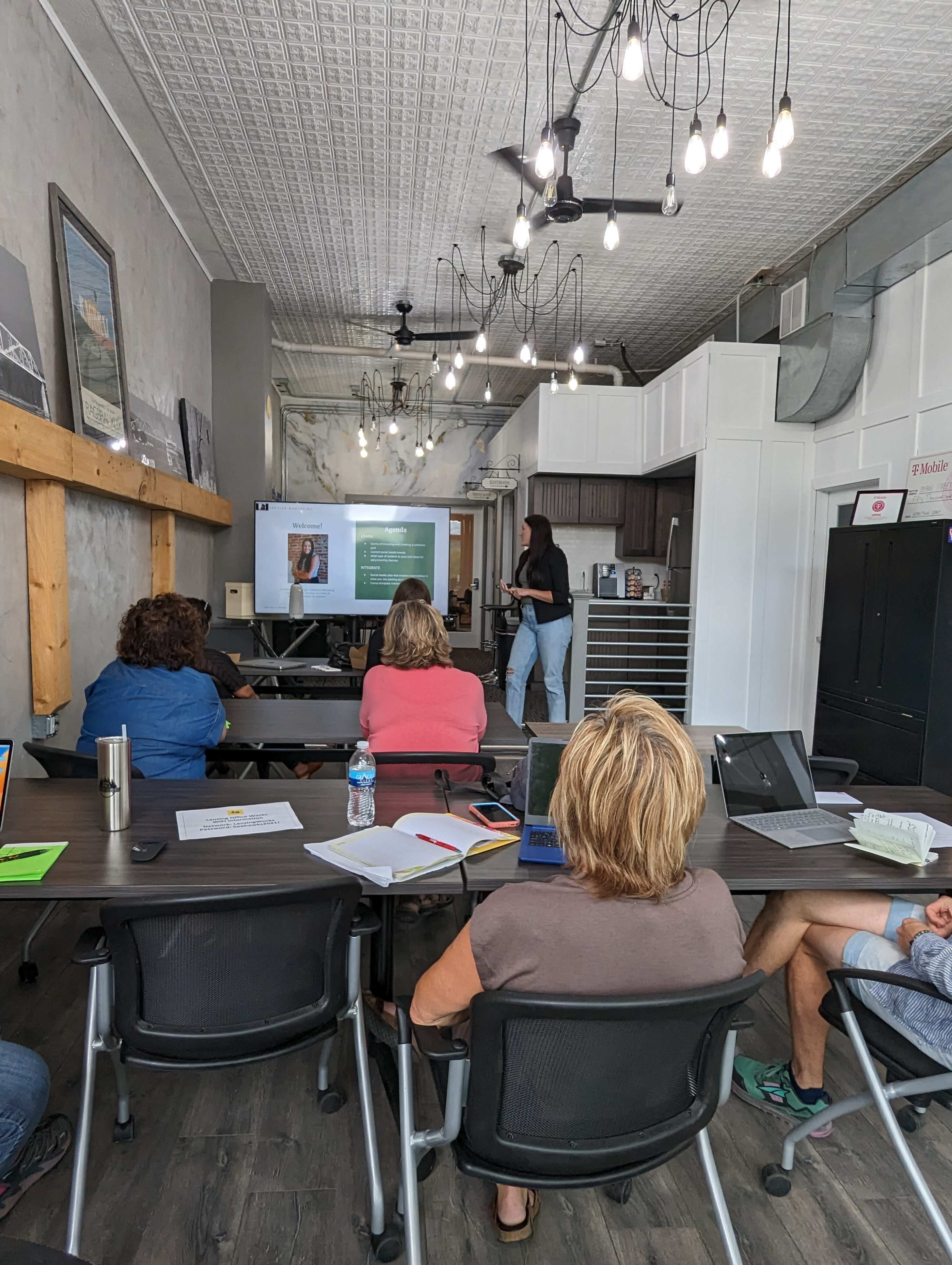 A group of people sit at tables in a bright room while a speaker presents information on a screen.