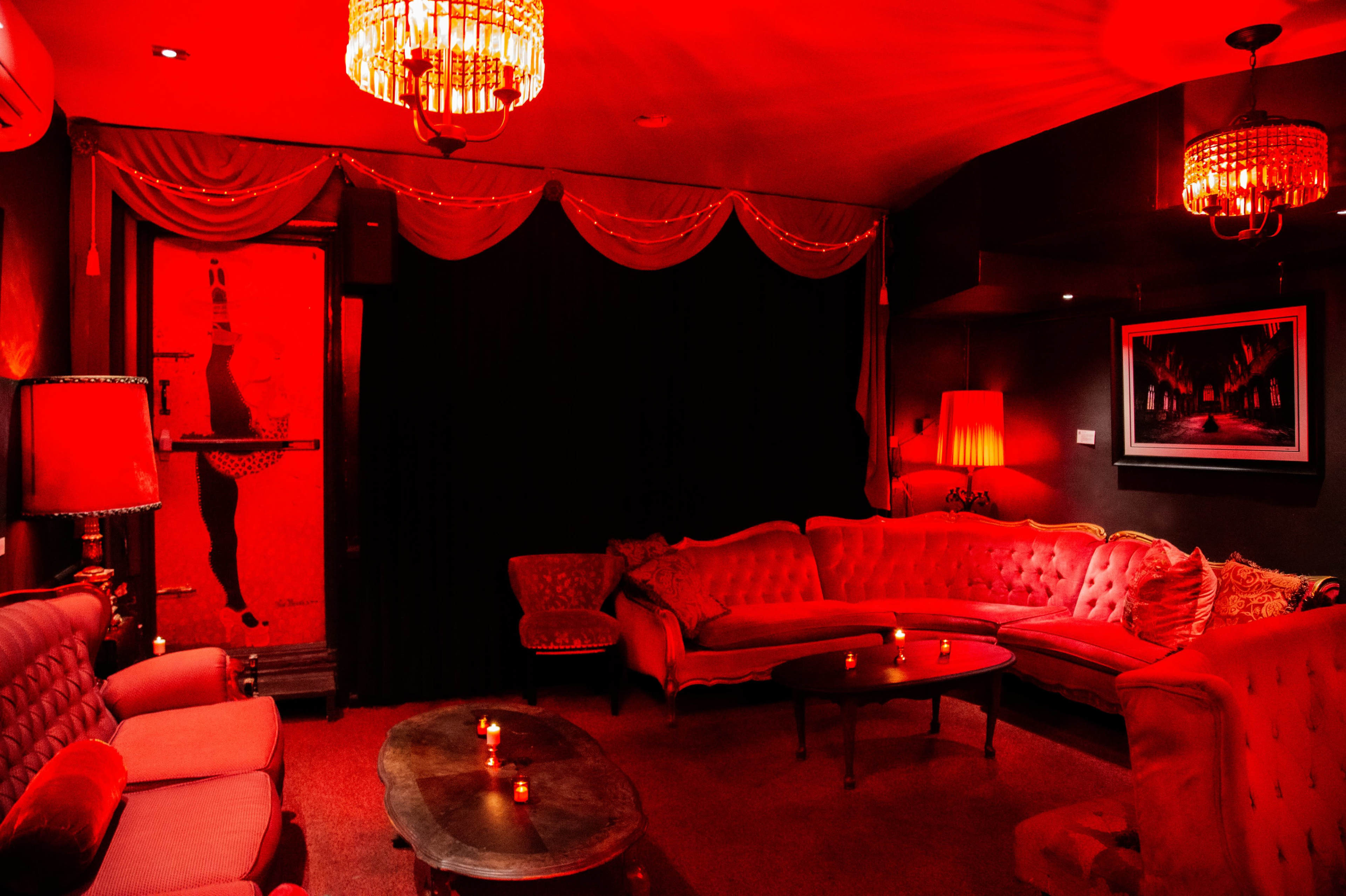 A dimly lit lounge features red decorative chandeliers, velvet sofas, and a wooden coffee table surrounded by soft candlelight.