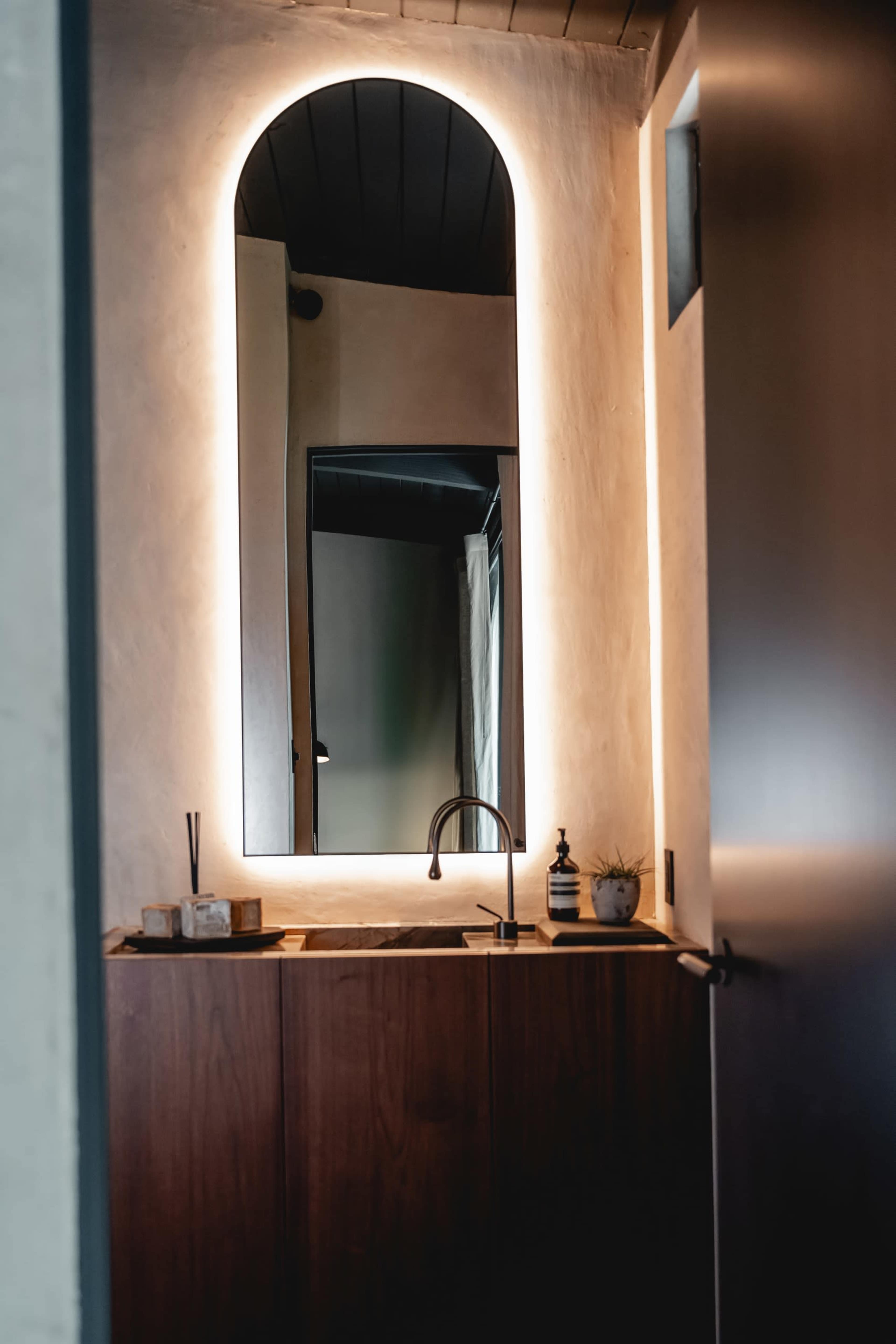 The image shows a modern bathroom with a wooden vanity, a mirror framed by ambient lighting, and a sleek faucet.