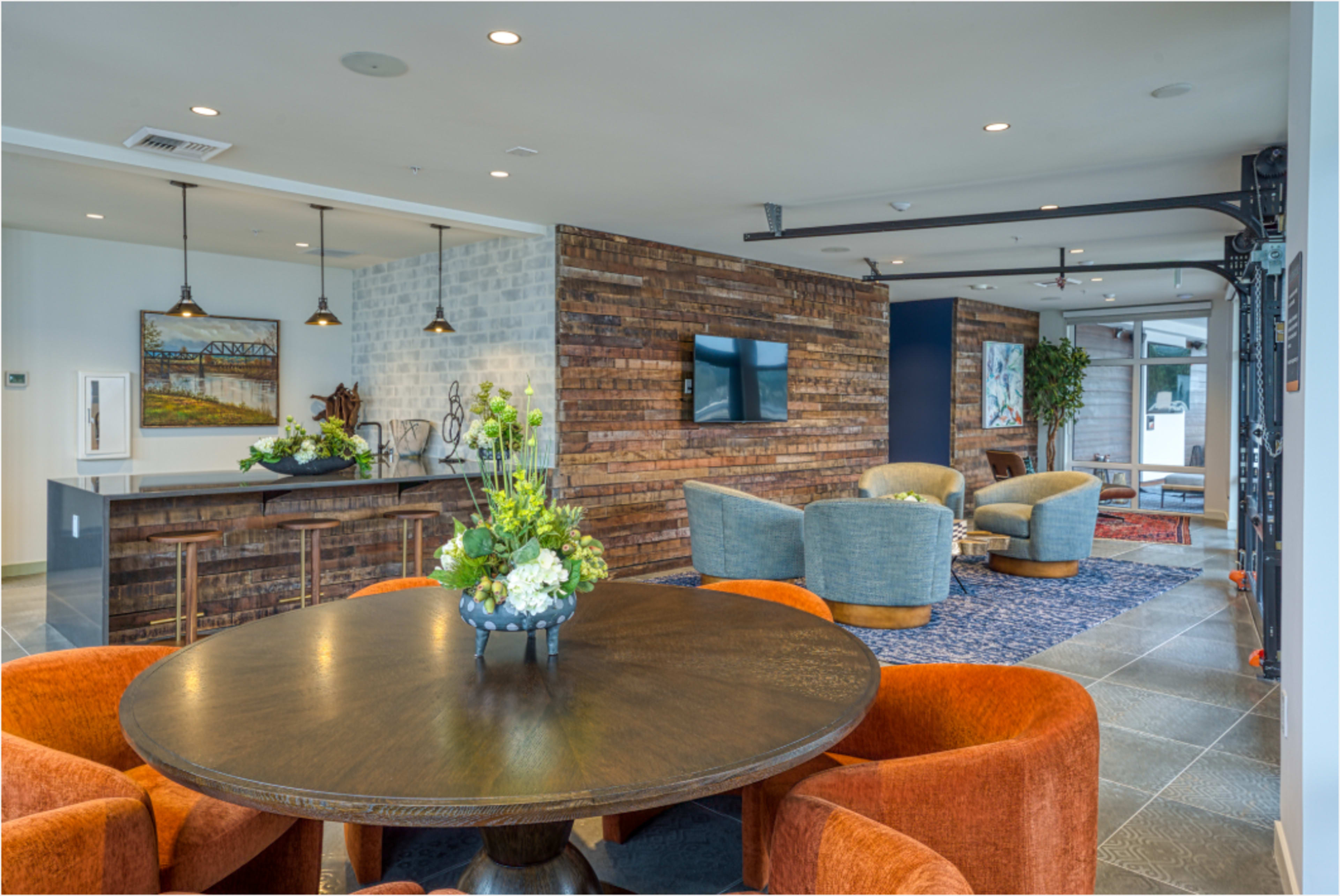 The image shows a modern lobby area featuring a round wooden table, upholstered chairs, and a reception desk against a backdrop of textured wood panels and a decorative painting.
