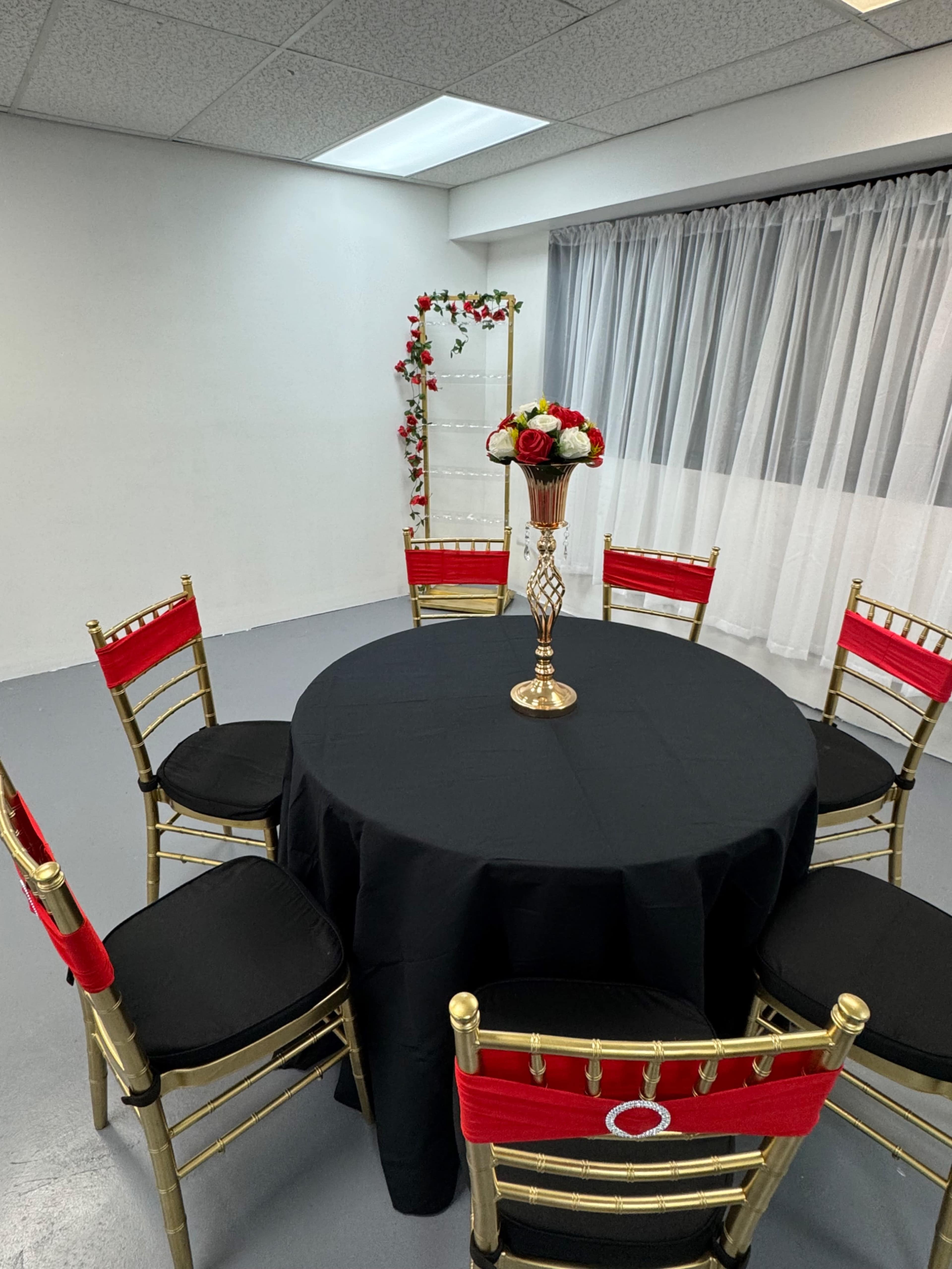A round black table is set with a golden vase of flowers, surrounded by five gold chairs with red seat cushions in a brightly lit room.