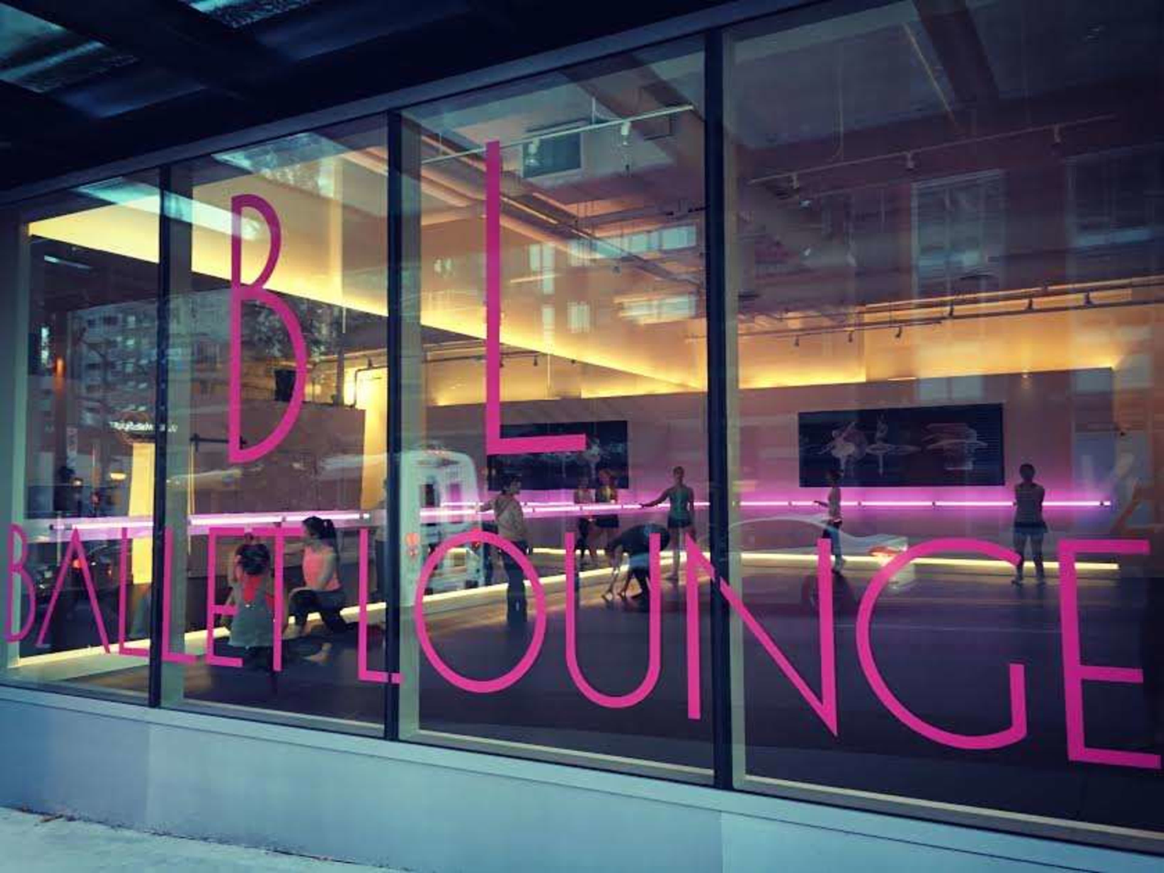 A dance studio with large glass windows displaying the words "Ballet Lounge" in bright pink lettering, where people are engaged in a ballet class inside.