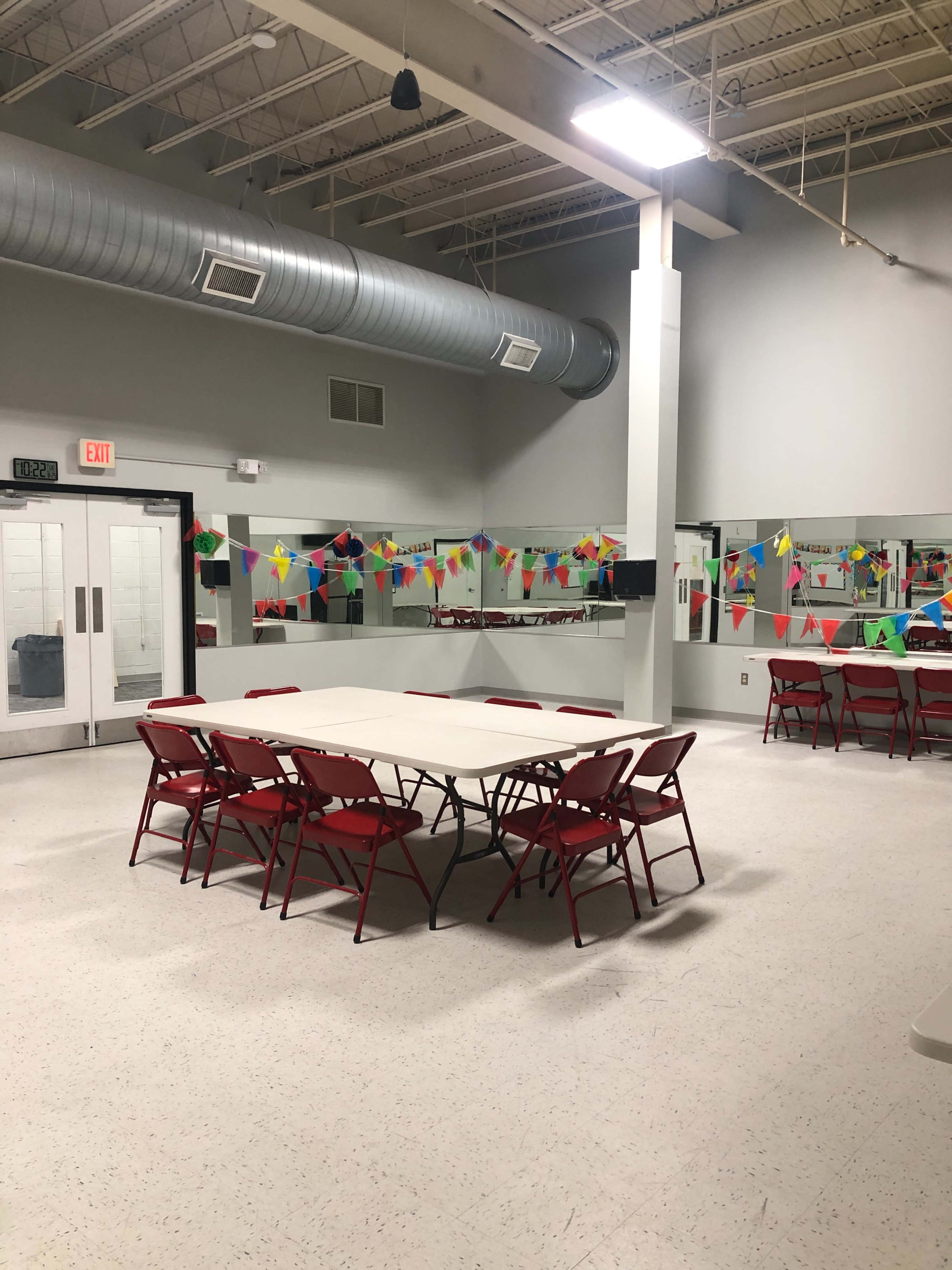 A large, empty room features a central folding table surrounded by red chairs, with colorful decorations along the walls and mirrors.