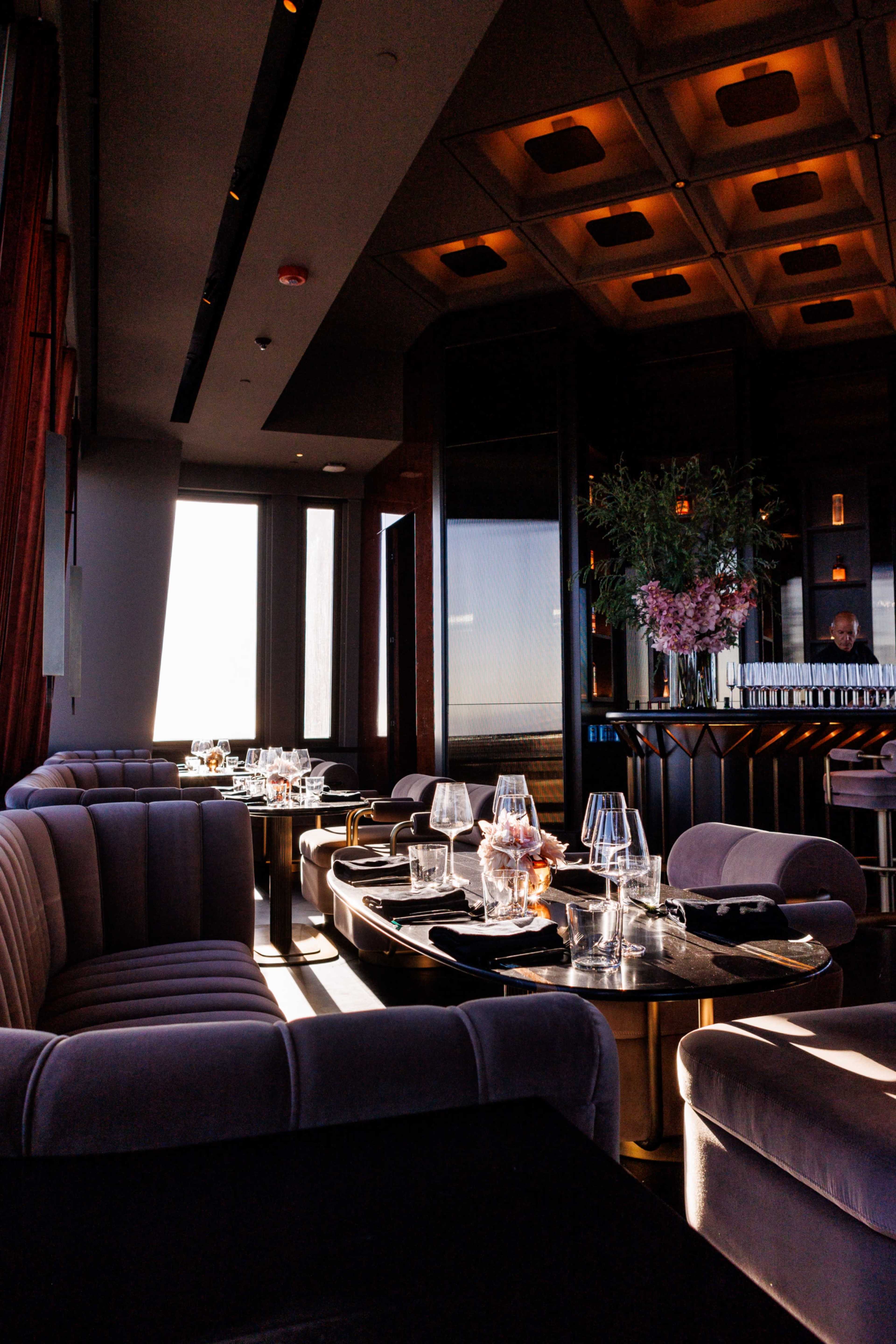 The image shows a modern lounge area with plush seating, a bar in the background, and large windows allowing natural light to fill the space.